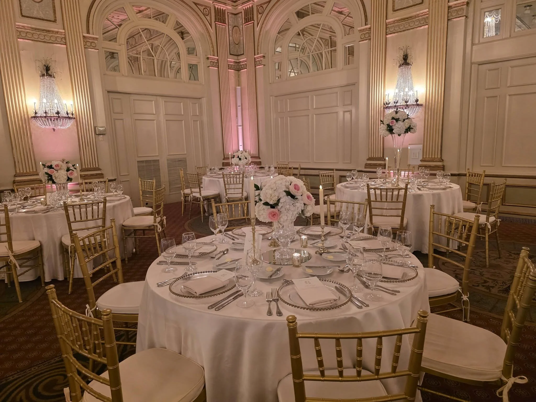 Elegant banquet hall with gold Chiavari chairs, round tables with white tablecloths, floral centerpieces of white and pink flowers, candles, and ornate chandeliers on the wall, in a room with high ceilings and detailed wall moldings.