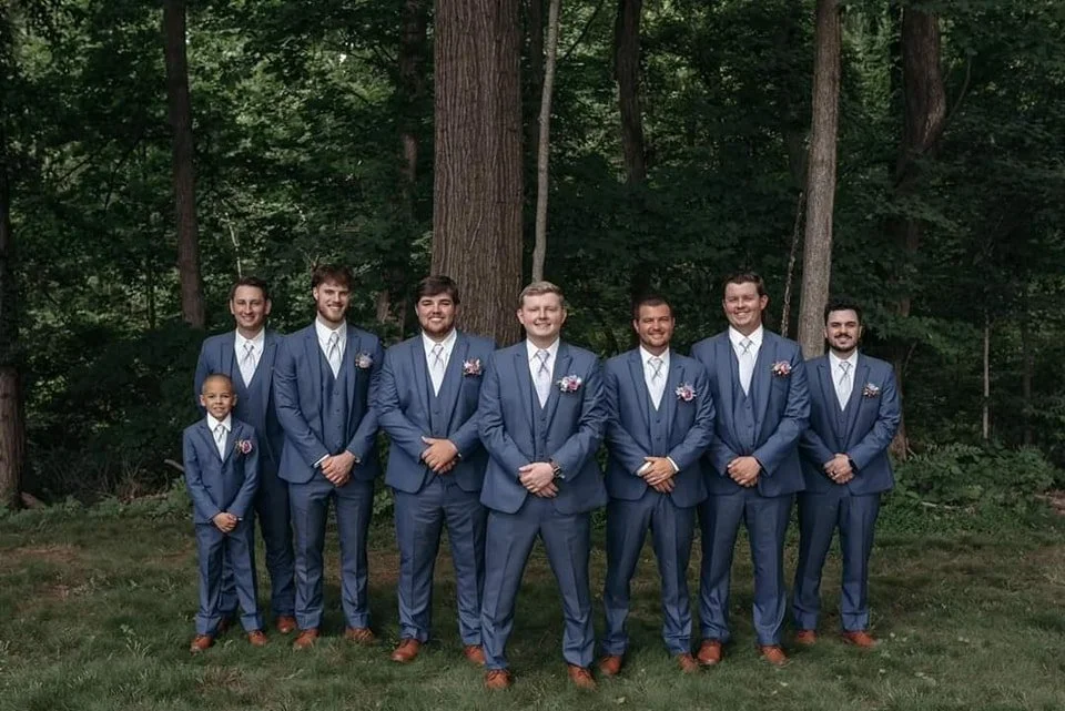 Group of men in matching blue suits, white shirts, and brown shoes standing outdoors in a wooded area, smiling at the camera.