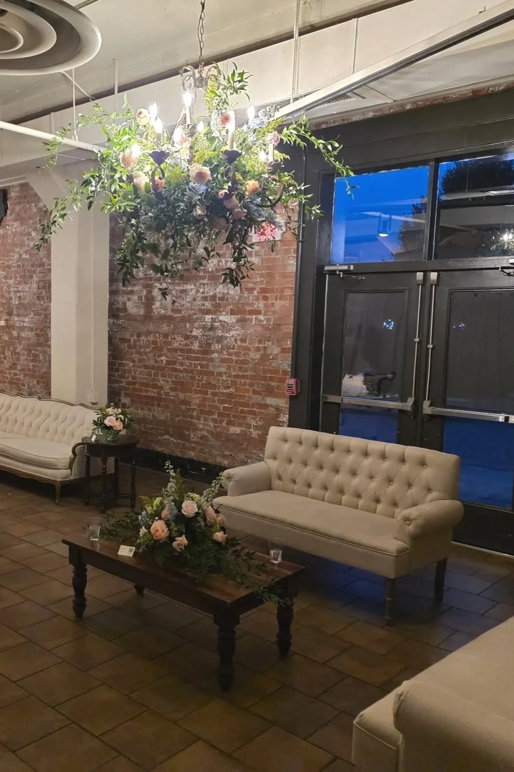 Living room with brick wall, beige tufted sofas, dark wooden coffee table with flowers, floral chandelier, large window with black frame, tiled floor.