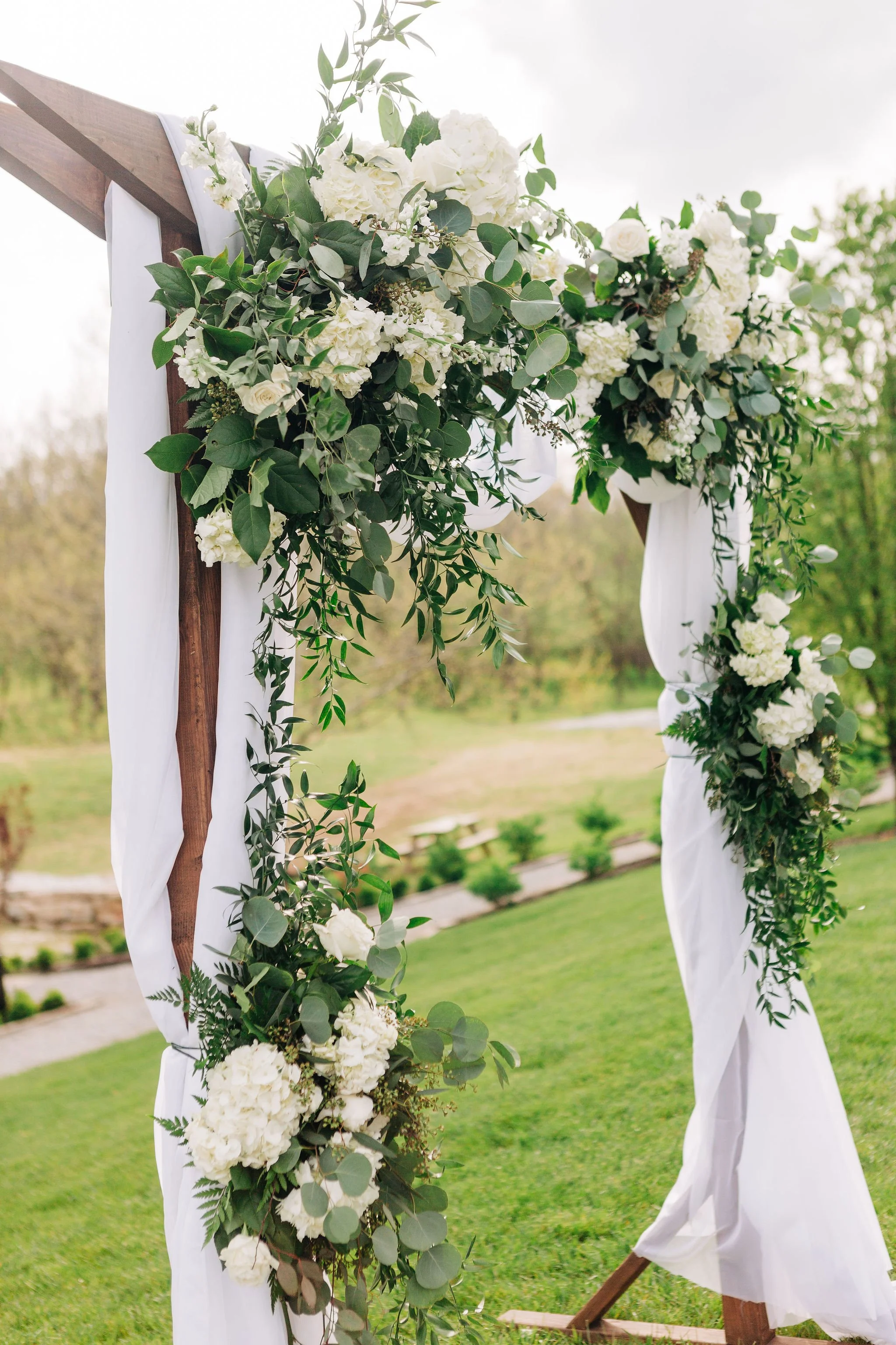 Close-up of a wooden wedding arch decorated with white flowers and greenery, draped with white fabric, outdoors on a grassy lawn.