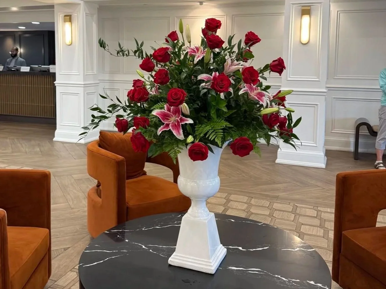 A large floral arrangement with red roses and pink lilies in a white urn, placed on a black marble table.