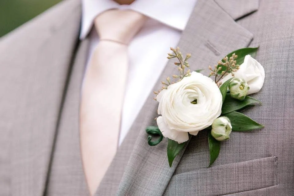 Close-up of a man's light gray suit with a white dress shirt and a cream-colored tie, featuring a white floral boutonniere on the left lapel.