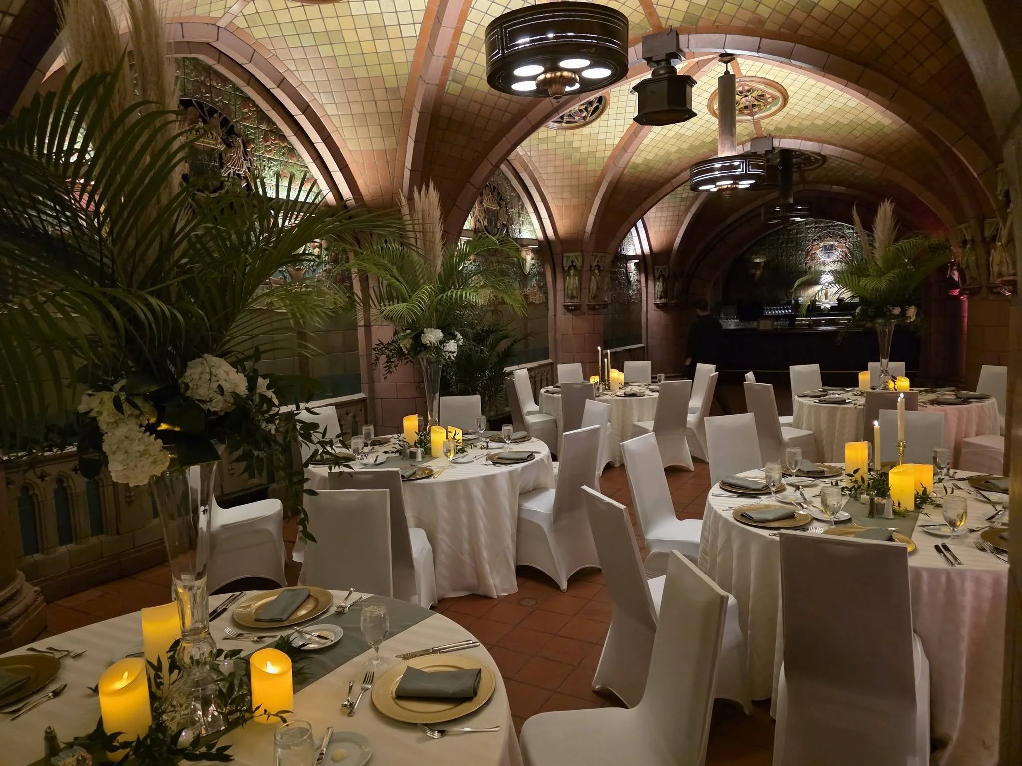 Elegant dining room with arched, stained-glass ceiling, decorated with plants and flowers, set with round tables, white chairs, candles, and tableware for a formal event.