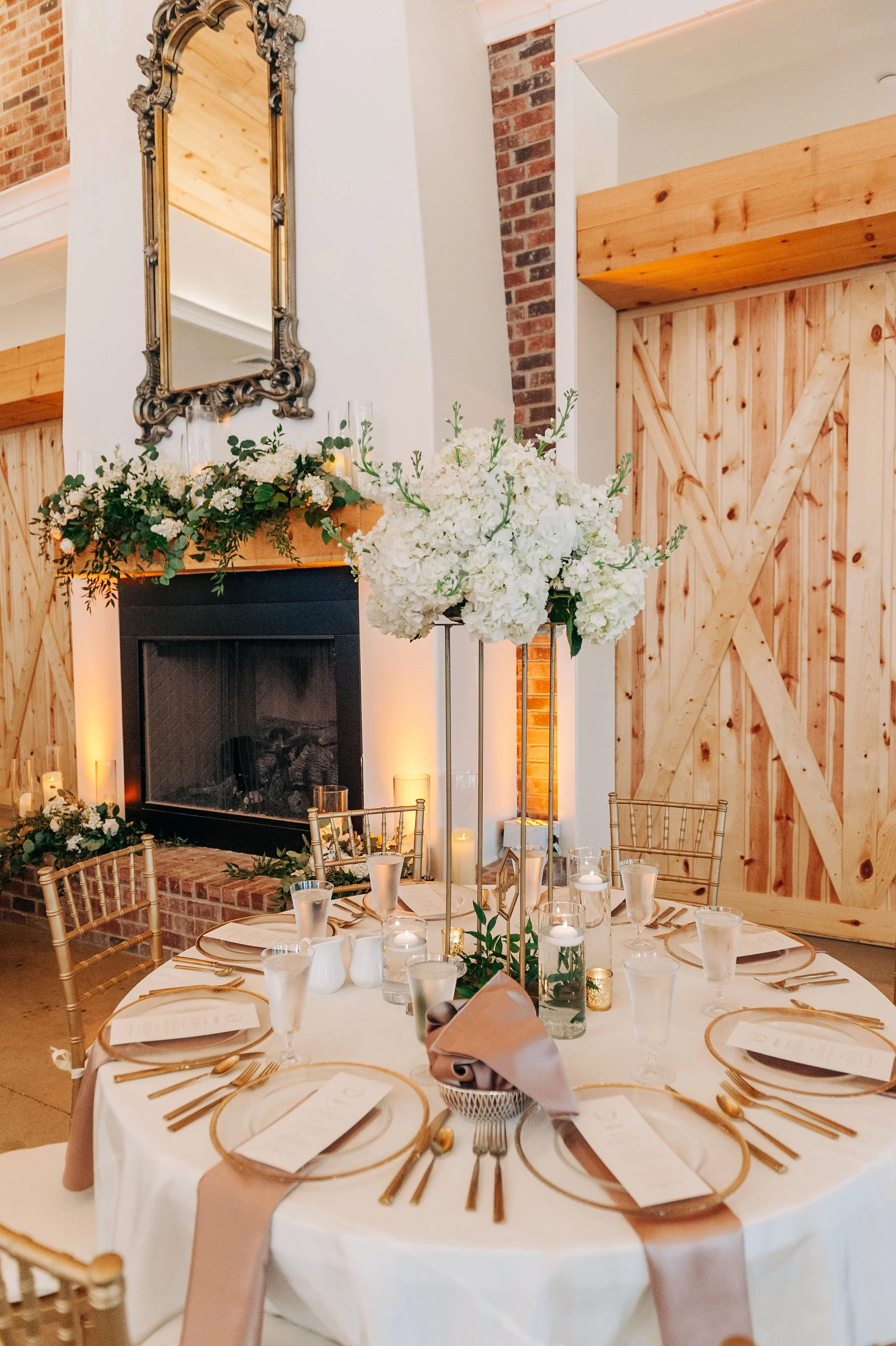 Round banquet table set with gold-cutlery, glasses, and menus. A tall floral centerpiece with white hydrangeas and greenery is in the middle, with candles around it. Wooden chairs surround the table. The background features a fireplace, wooden wall, and a large ornate mirror above the mantel decorated with white flowers and candles.