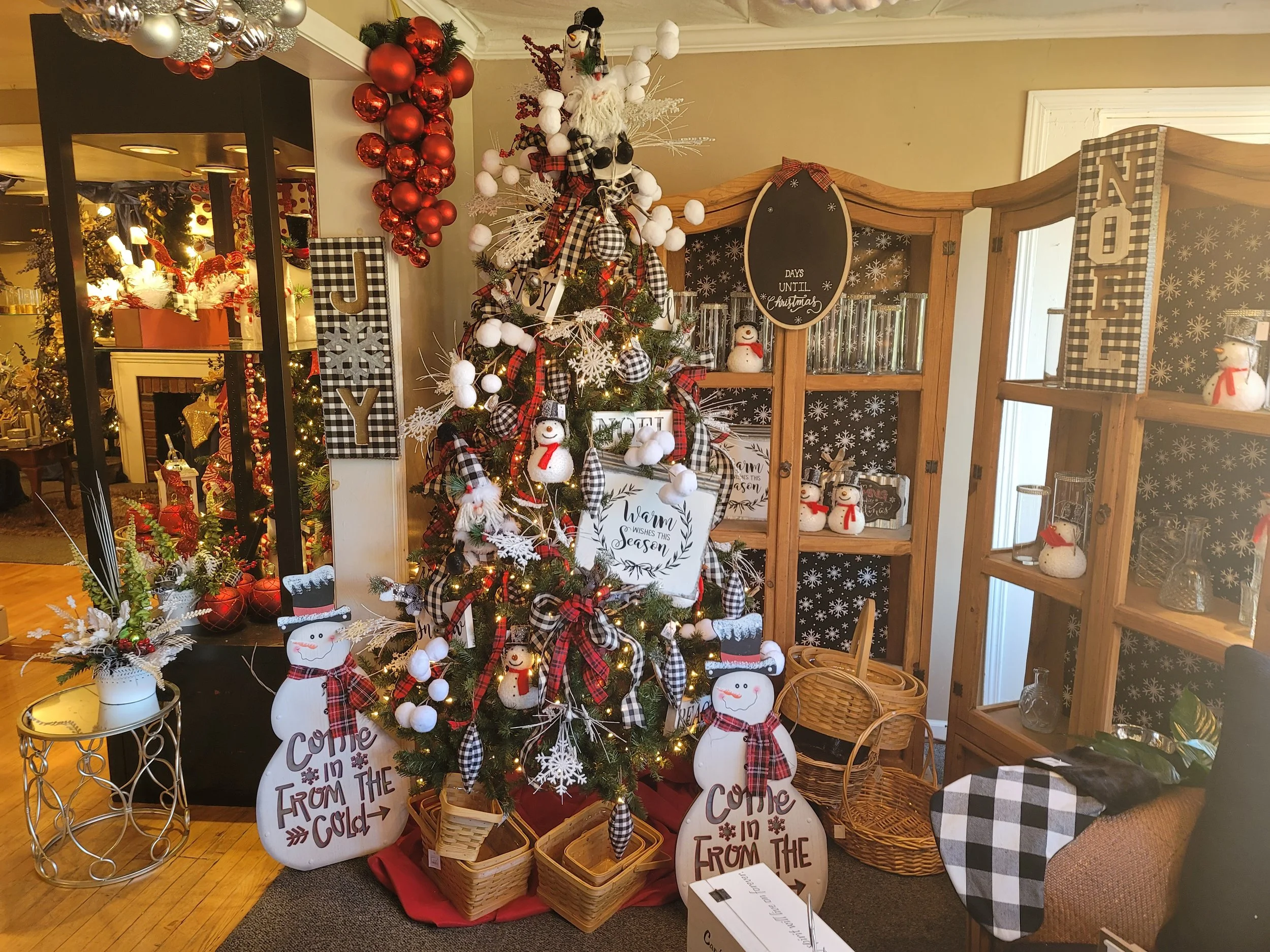 A decorated Christmas tree with snowman and snowflake ornaments, surrounded by signs saying 'Come in from the cold,' 'Warm wishes this season,' and 'Joy.' Red and black checkered ribbon, red baubles, and white cotton ball decorations adorn the tree. In the background, there are shelves with snowman decorations, a black chalkboard sign showing 'Days until Christmas,' and other holiday decor.