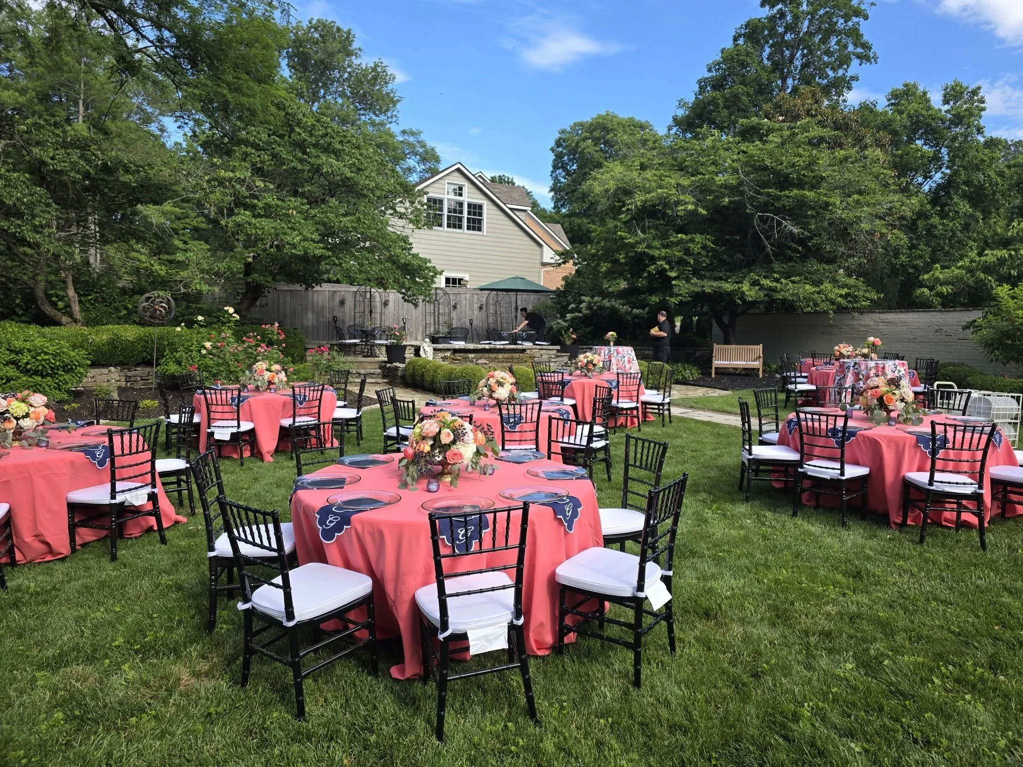 An outdoor backyard setup for a celebration with round tables covered in pink tablecloths, floral centerpieces, black chairs, and a patio area with two people setting up. The yard has lush green grass, trees, a wooden fence, and a house in the background under a blue sky.