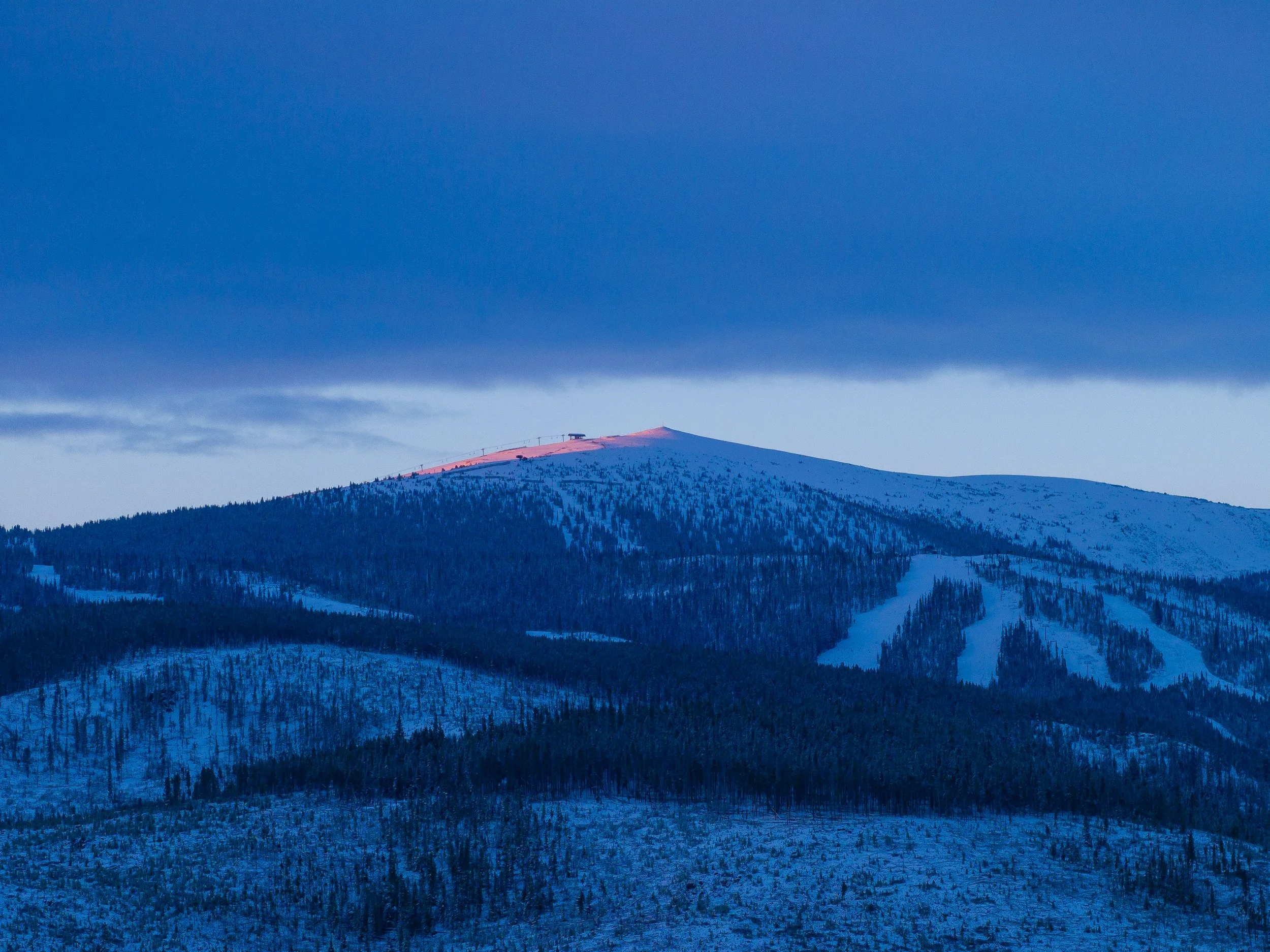 Snow-covered mountain under a dark, cloudy sky with the setting or rising sun casting a pink glow on the peak.