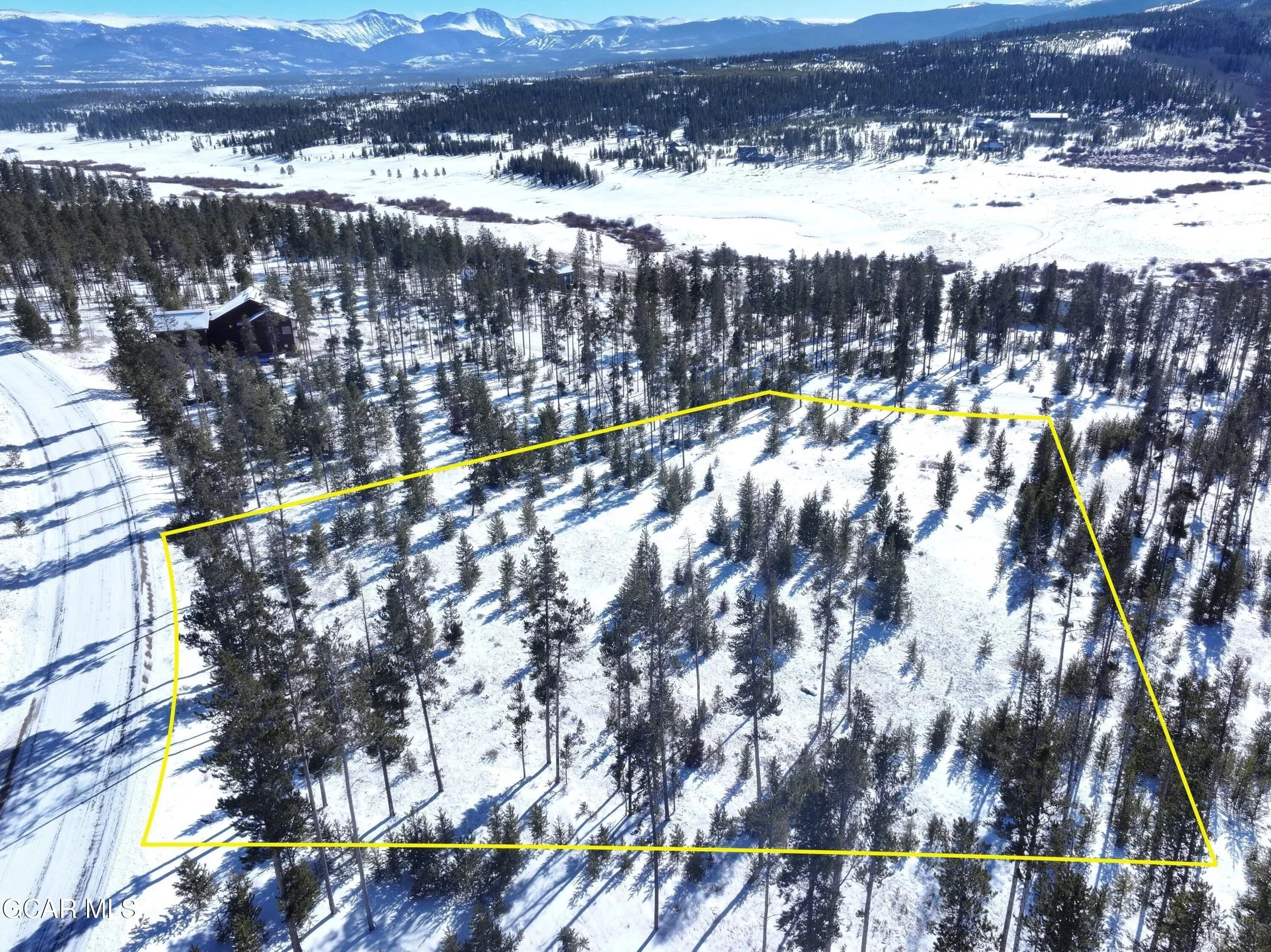 A snowy landscape with a forest of pine trees, mountains in the background, and a few houses scattered across the terrain. A yellow line outlines a section of the forested area.