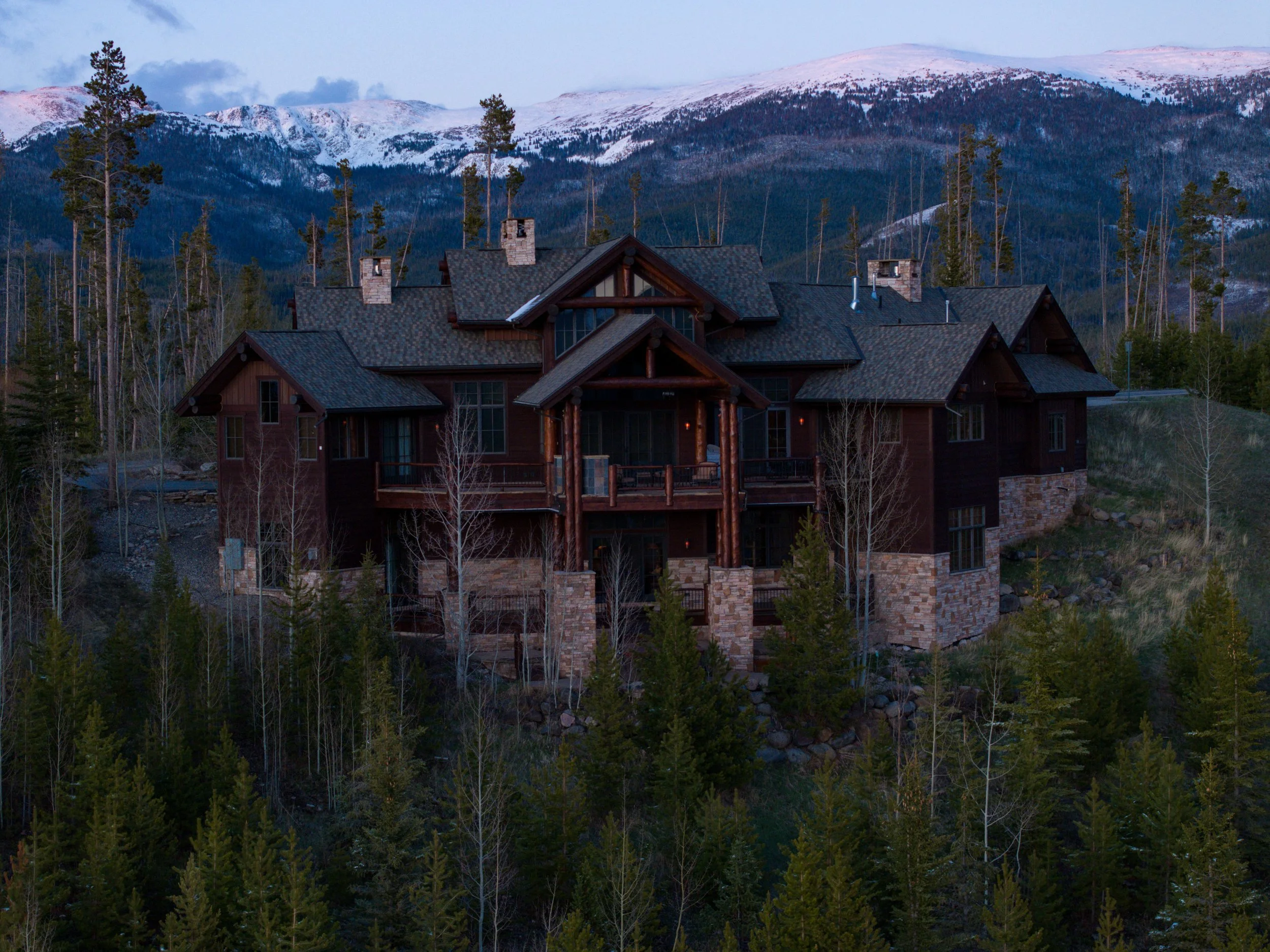A large wooden mountain house surrounded by trees with snow-capped mountains in the background.