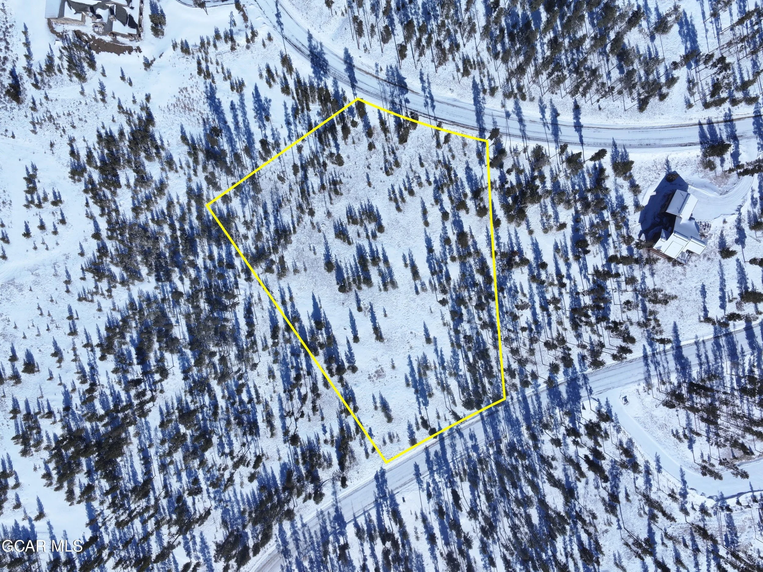 A snowy aerial view of a wooded area with a yellow outlined property. There are trees, roads, and two houses visible at the edges of the image.