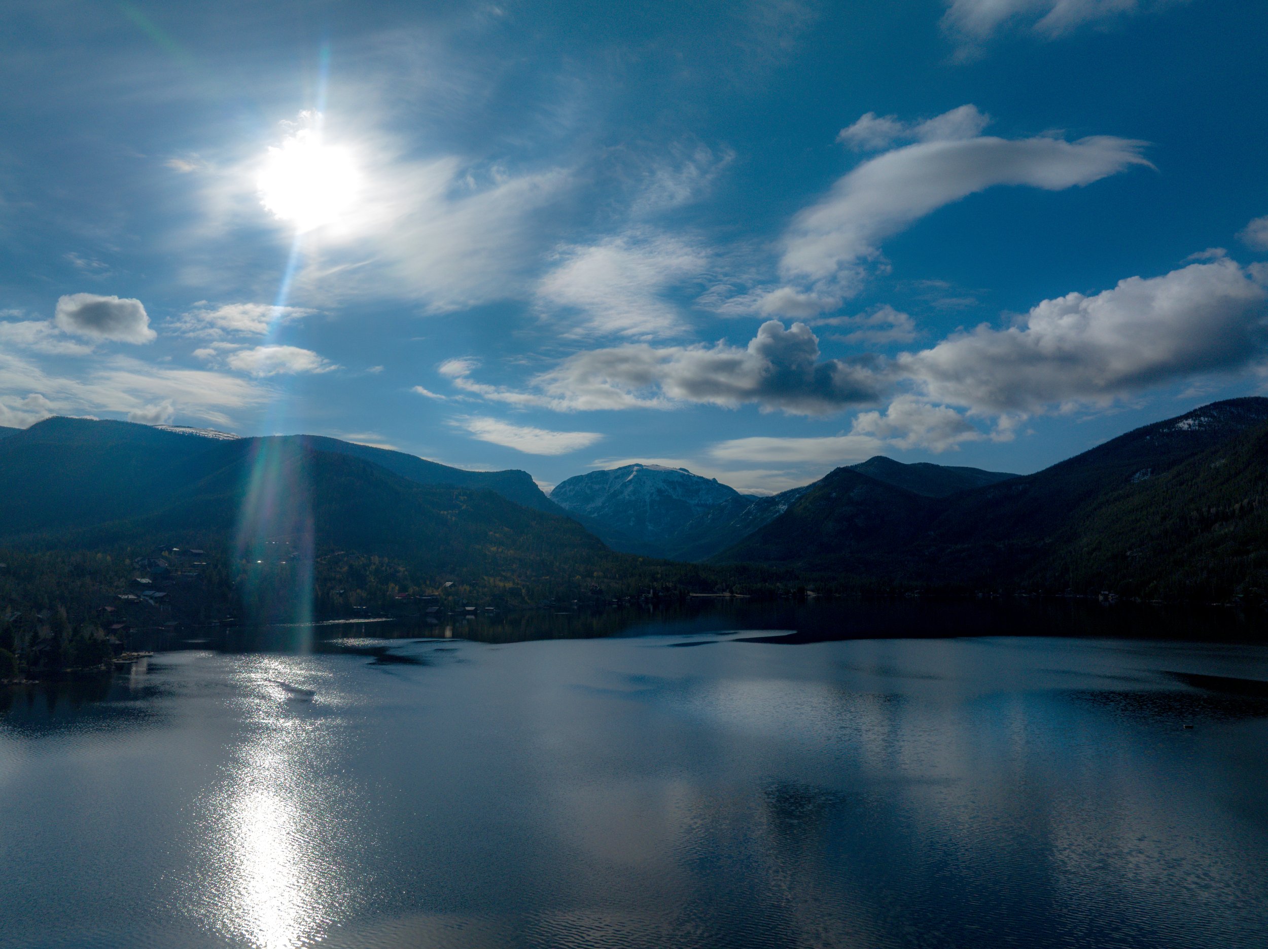 A scenic view of a blue lake surrounded by mountains under a bright sky with scattered clouds and the sun shining.