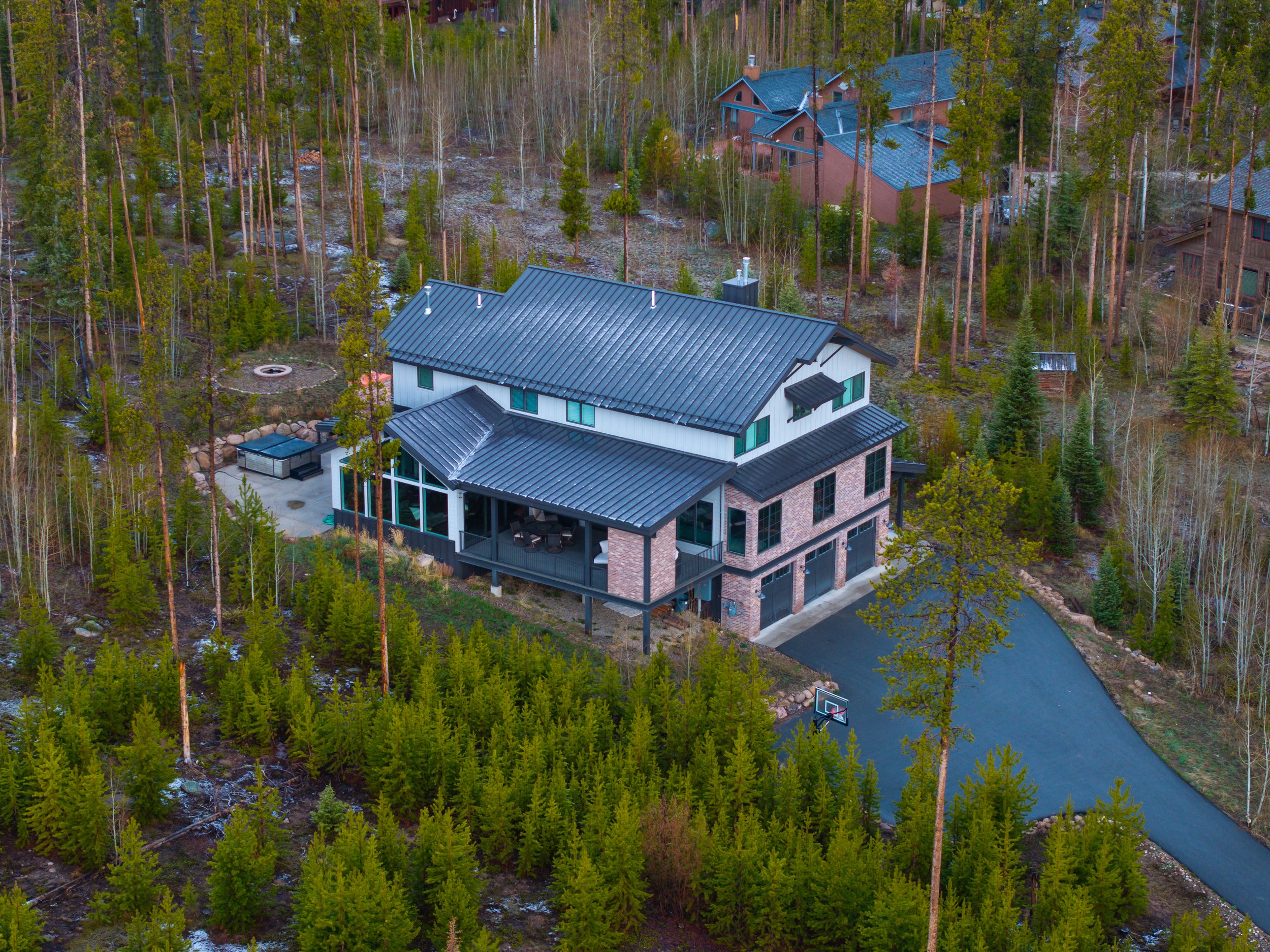 Aerial view of a modern house with a metal roof, surrounded by a forest with tall trees, a driveway, a small basketball court, and a backyard with outdoor seating and a fire pit.