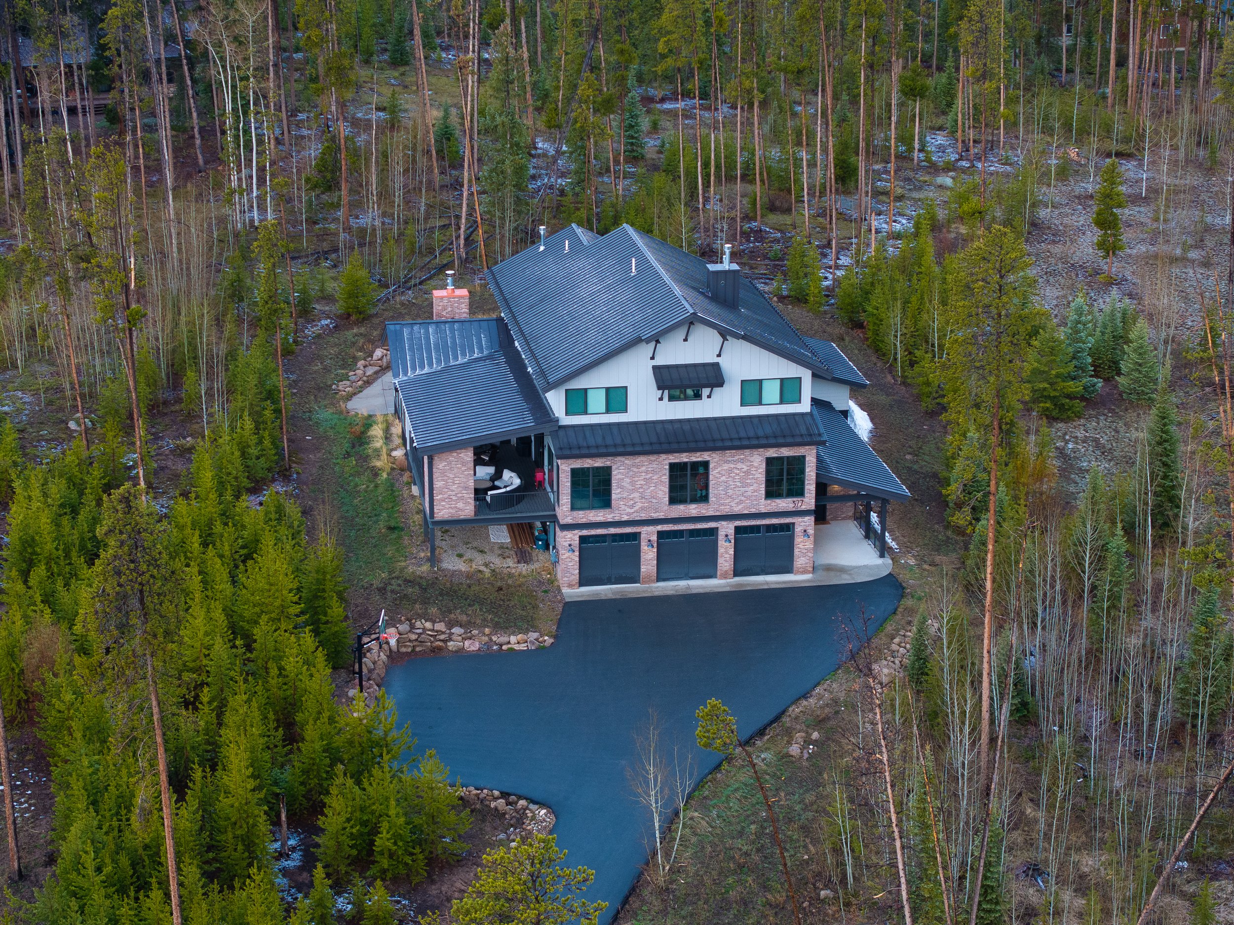 A house with a black roof and brick exterior situated in a wooded area with tall trees, a driveway, and a pond.