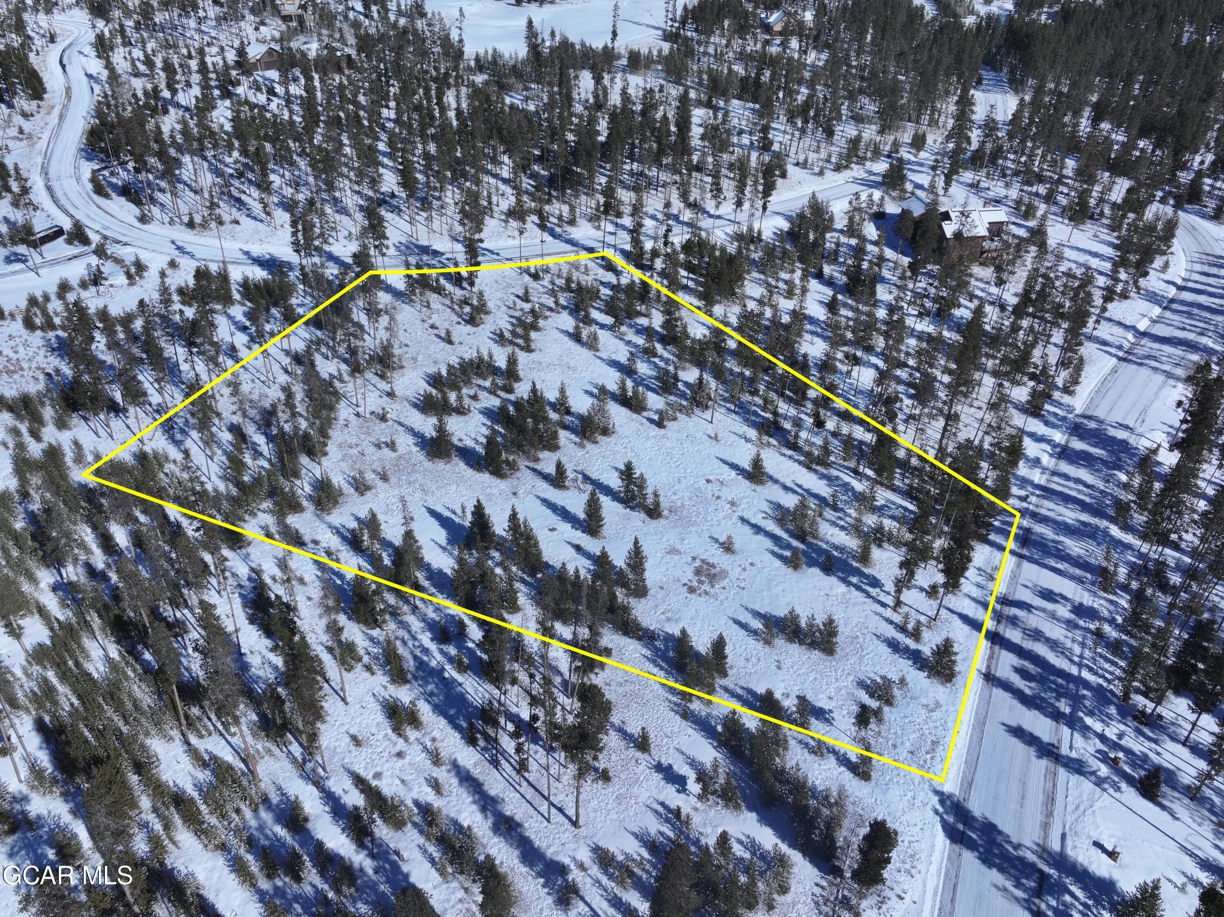 A snow-covered wooded lot outlined with yellow lines, with a road on its side and houses in the background.