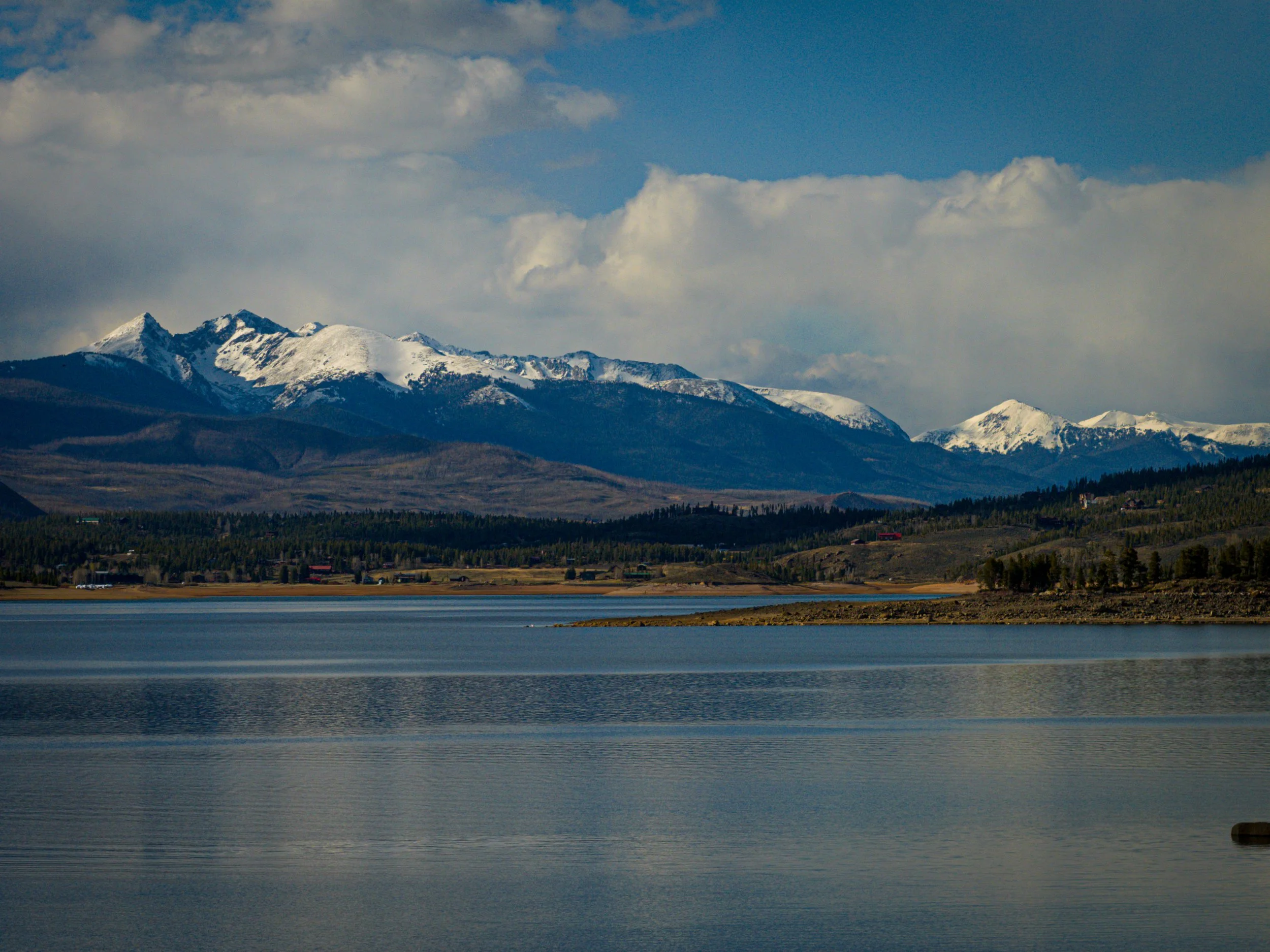 Snow-capped mountains beneath a partly cloudy sky, with a body of water in the foreground.
