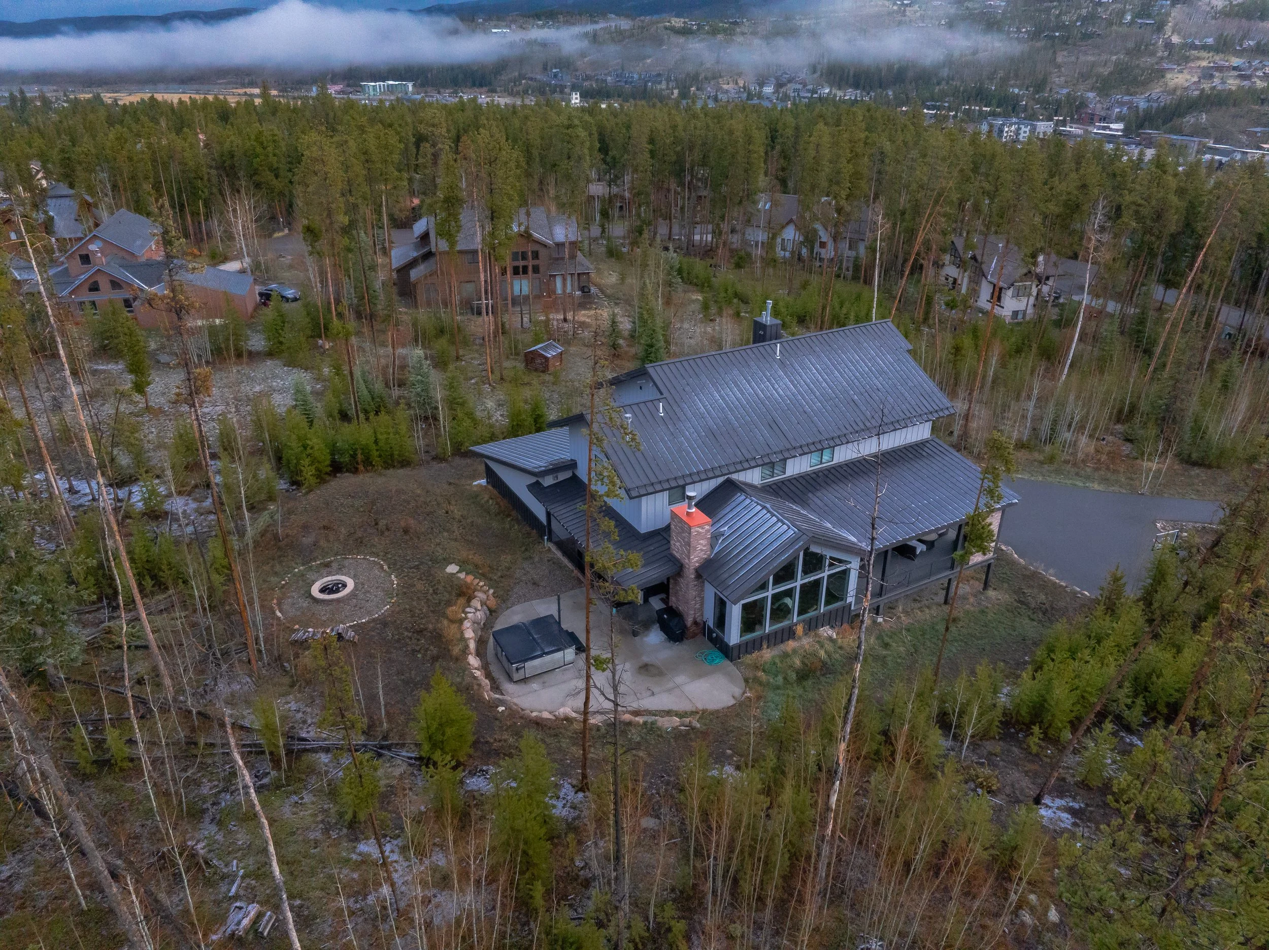 Aerial view of a residential area with houses surrounded by tall pine trees, some rooftops are visible, and mist or low clouds are drifting over the distant hills.
