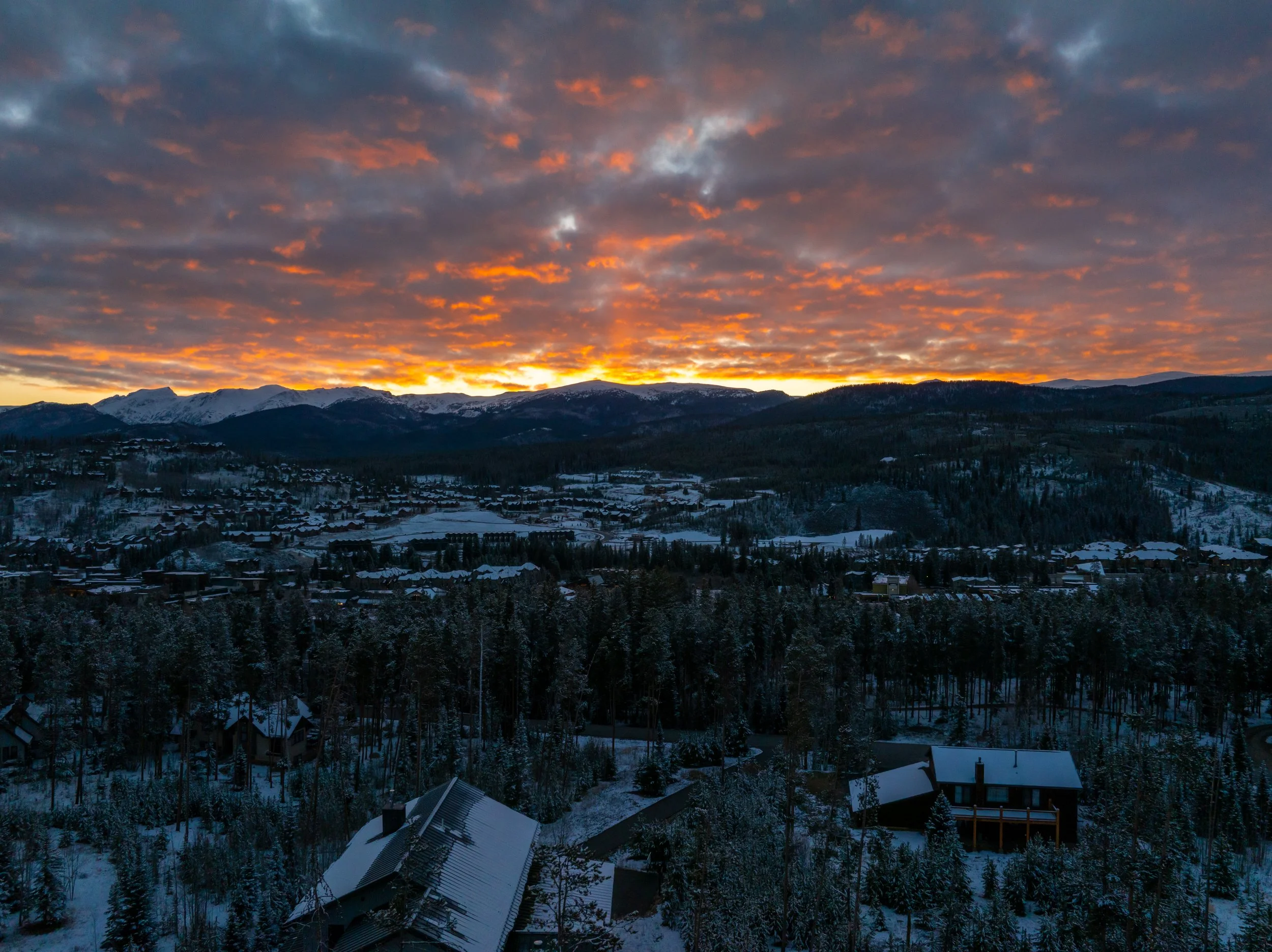 A scenic view of a mountain range at sunset with dark clouds and an orange and yellow sky, overlooking a snow-covered town with houses and trees.