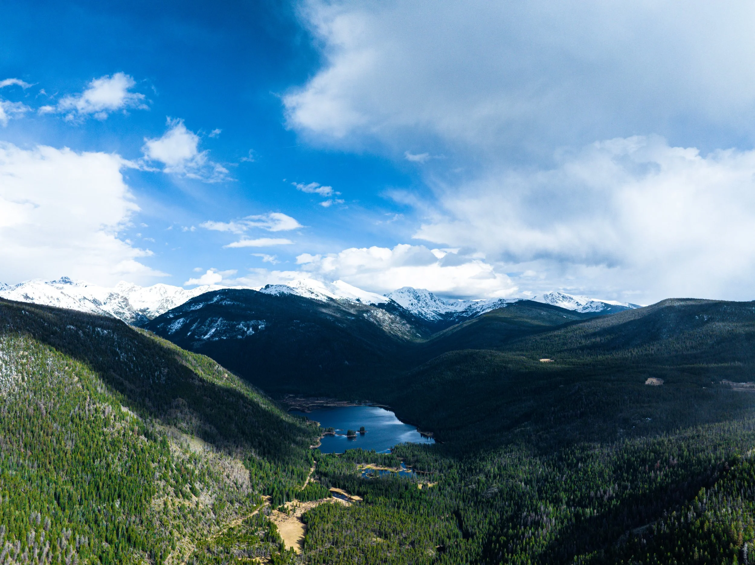 A scenic landscape of snow-capped mountains, dense green forests, a lake, and a partly cloudy sky.