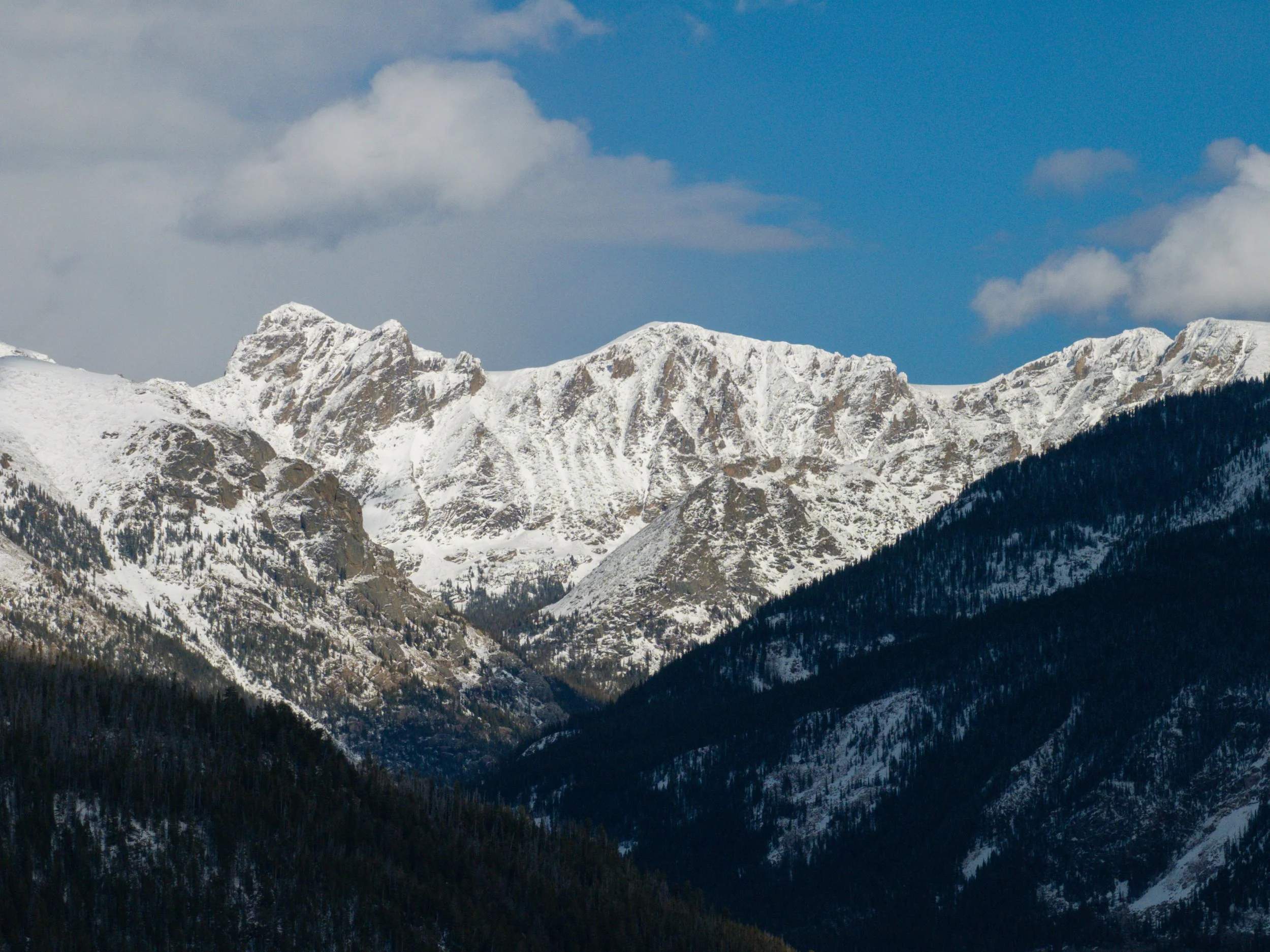 Snow-covered mountain peaks under a partly cloudy sky with some trees at the mountain base.