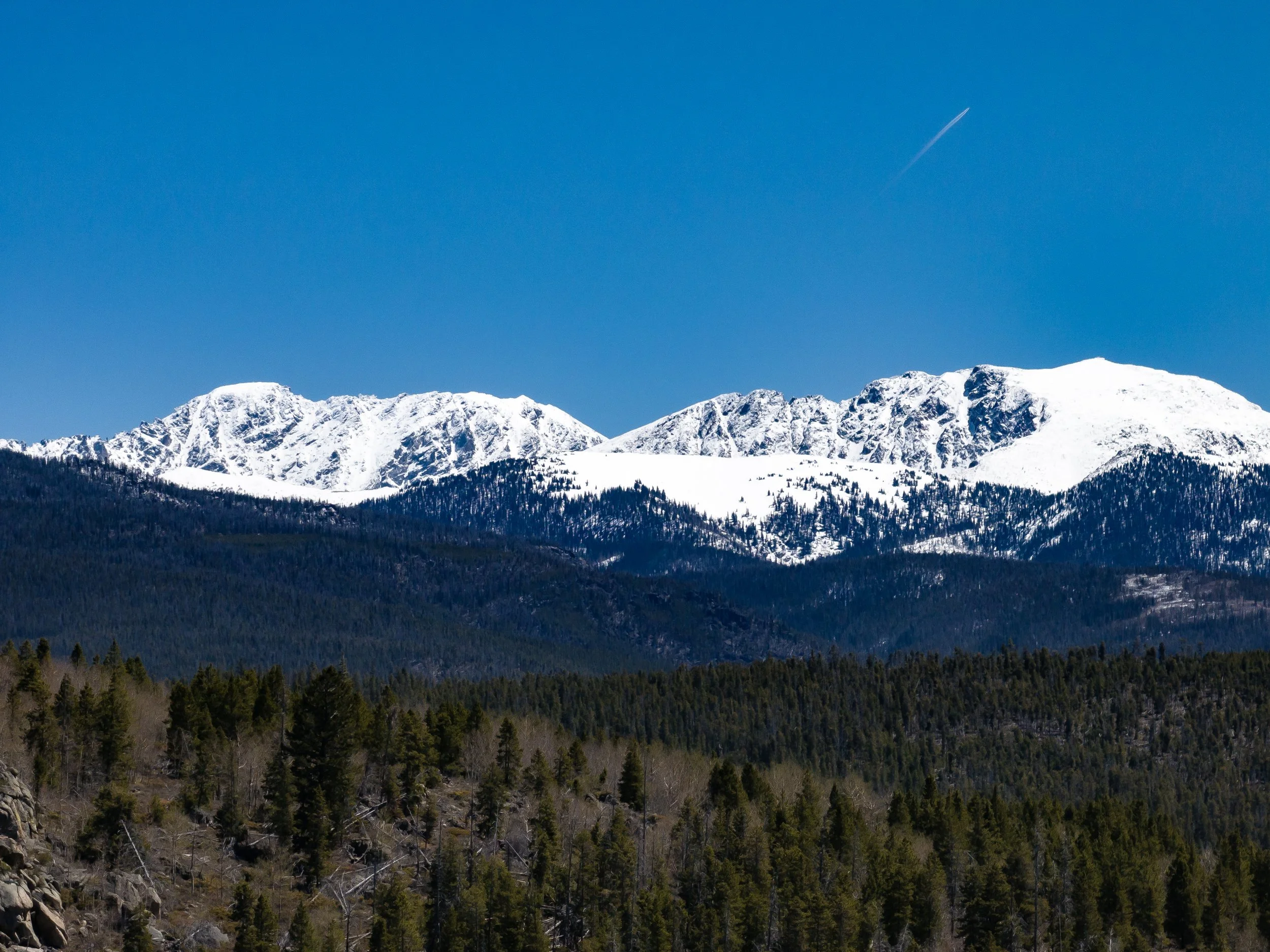 Snow-capped mountain range under a clear blue sky with a distant airplane contrail.