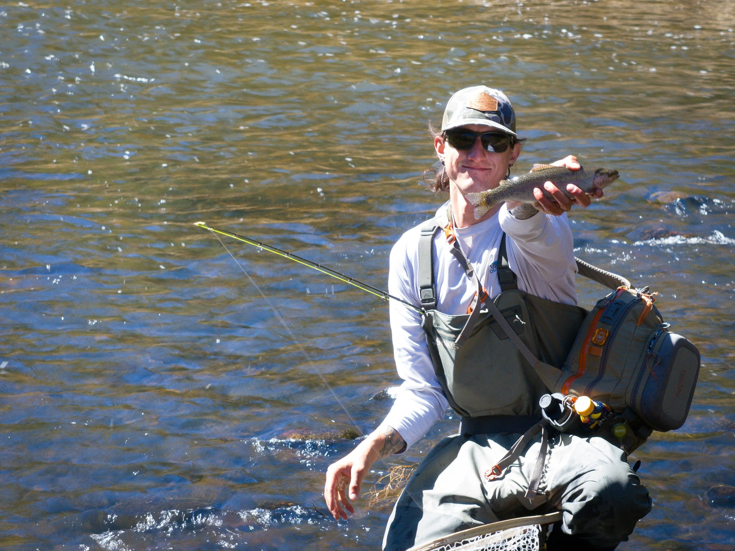 A person wearing sunglasses, a camouflage cap, and fishing gear holding up a fish near a body of water.