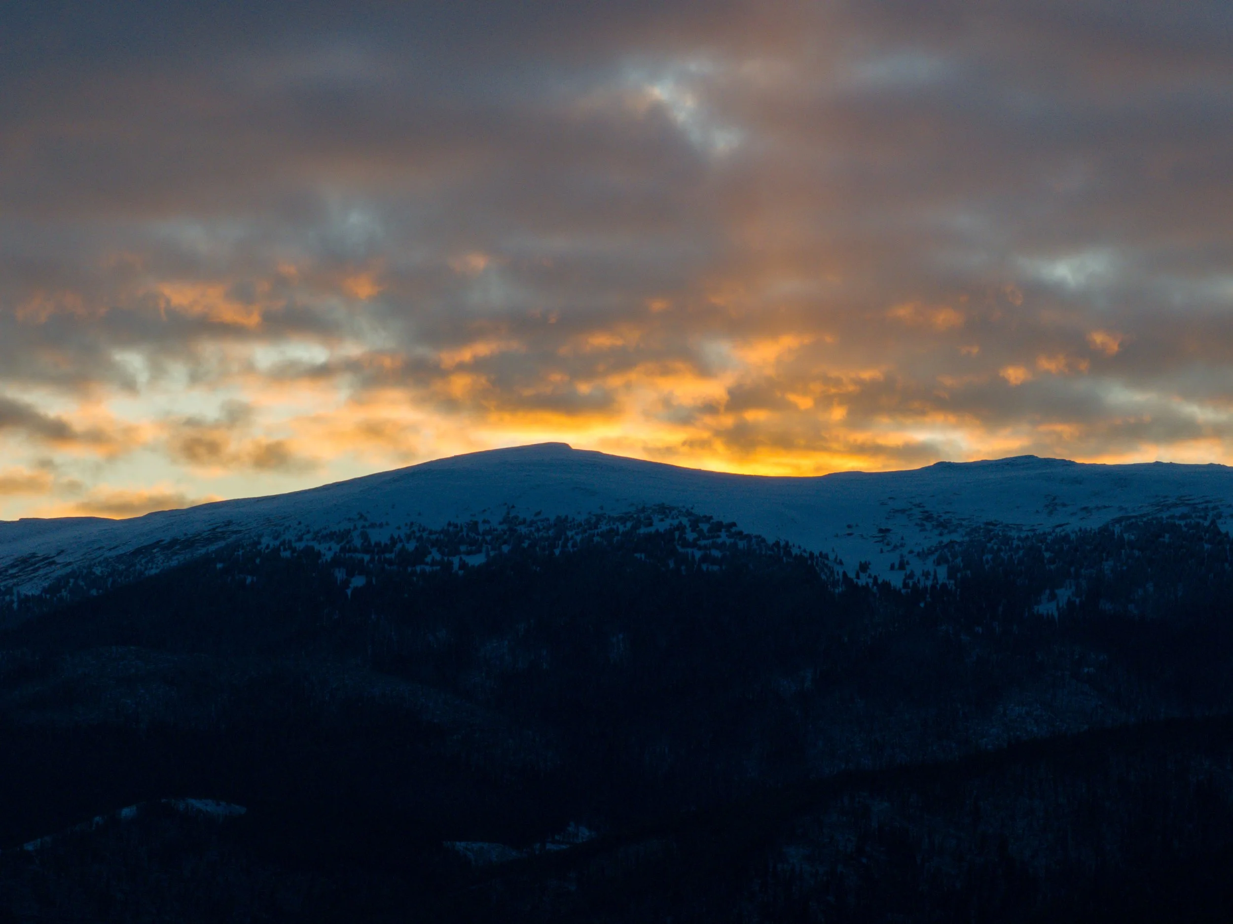Snow-covered mountain landscape at sunset with a partly cloudy sky illuminated in orange and yellow hues.