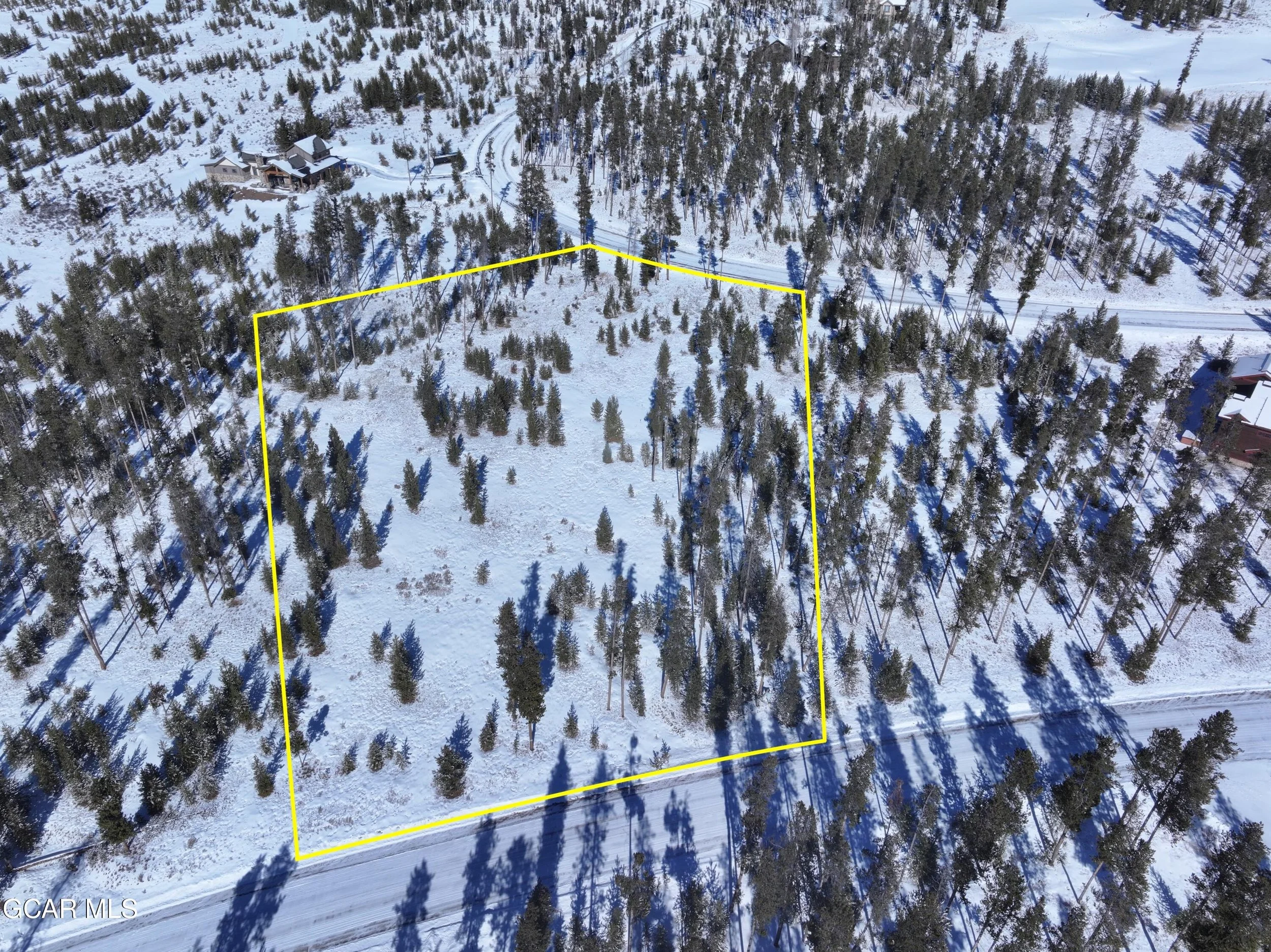 Aerial view of a snow-covered wooded lot surrounded by trees, with a yellow outline marking the property boundaries and a road at the bottom of the image.