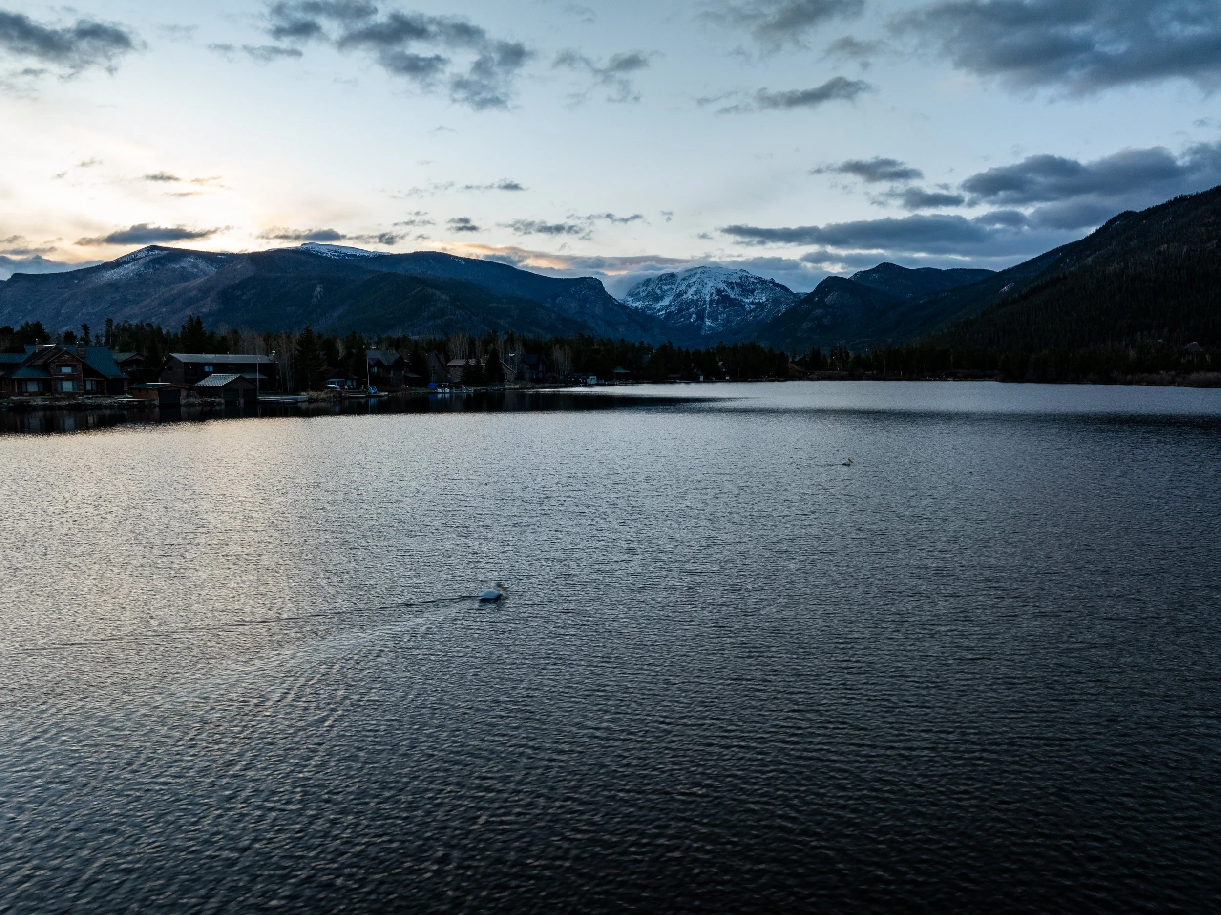 A body of water with ripples, surrounded by mountains with some snow on their peaks, and a sky with dark clouds and a hint of light near the horizon.