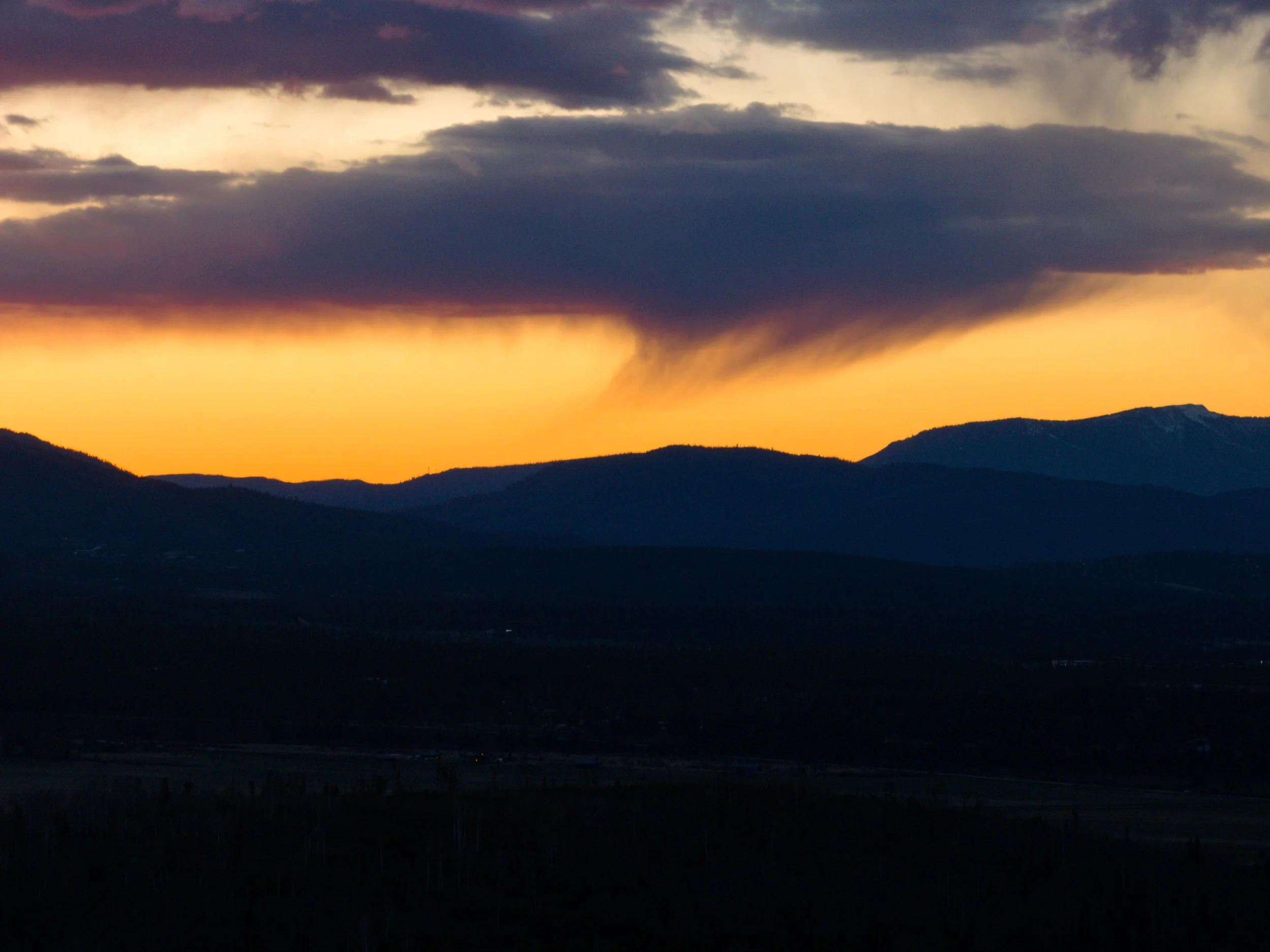A landscape view of mountains during sunset with a colorful sky featuring shades of purple, orange, and yellow, and some dark clouds.