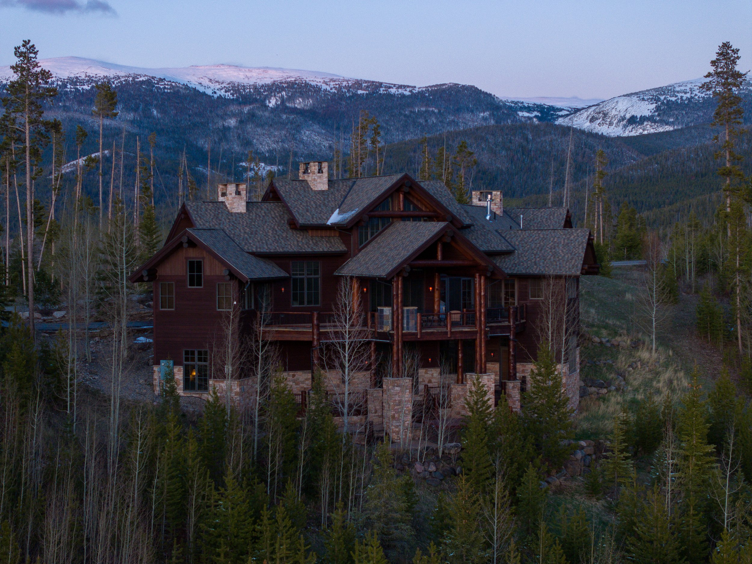 A large wooden house with multiple chimneys, nestled among tall pine trees, with snow-capped mountains in the background during early evening or dawn.