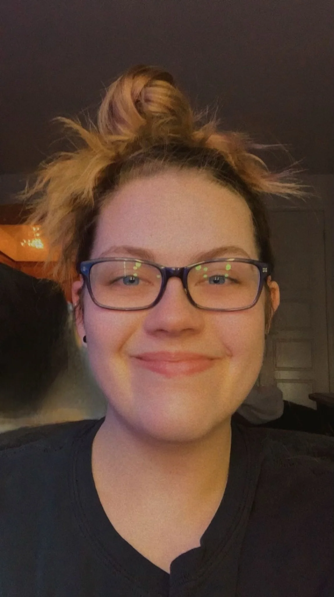 A woman with glasses, light skin, and reddish-brown hair styled in a messy bun, smiling at the camera in an indoor setting.