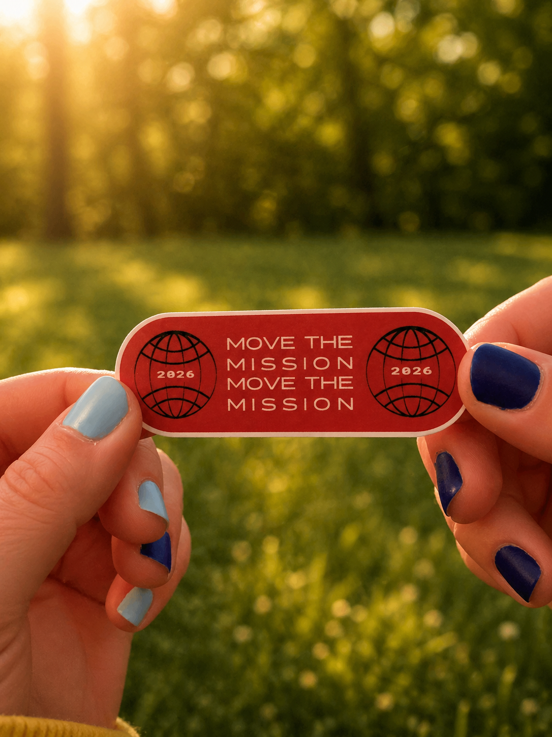 A person with blue and light blue painted nails is holding a small rectangular sticker with a red background, featuring two globe icons, the year 2026, and the phrase 'Move the Mission' repeated twice, outdoors with sunlight filtering through trees.