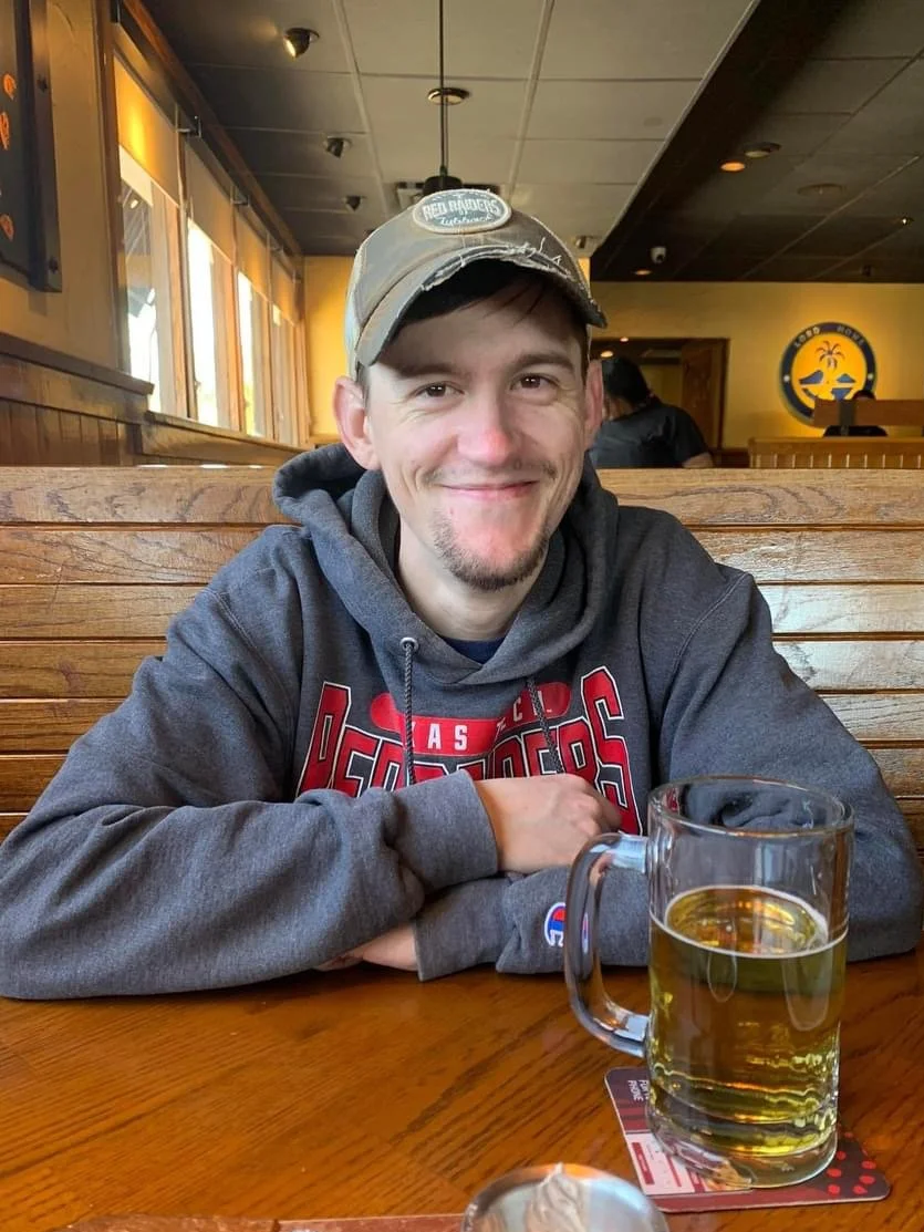 A smiling man wearing a baseball cap and gray hoodie sitting at a wooden table with a mug of beer in a restaurant or bar.