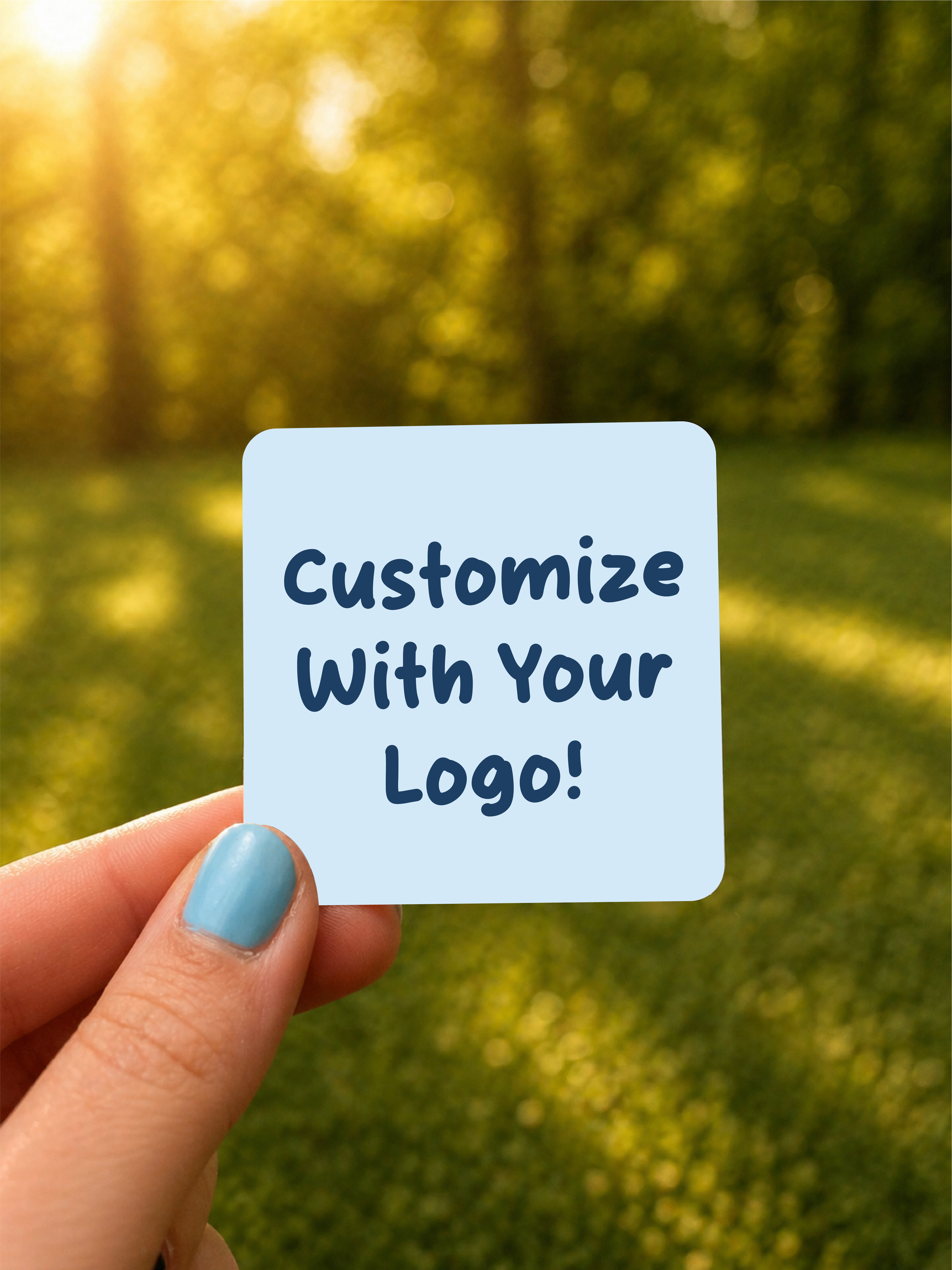 A hand with painted nails holding a light blue sticker that says "Customize with Your Logo!" against a blurred outdoor background with greenery and sunlight.