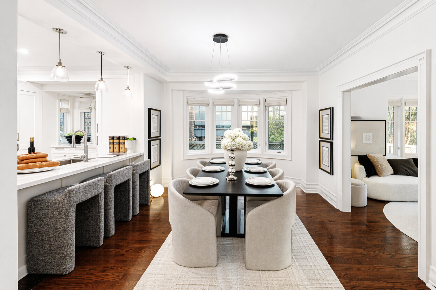 Bright dining room with a black table, beige chairs, and a white vase with white flowers, next to a kitchen with a white marble countertop and gray barstools, and a living room with a white sofa visible through a doorway.