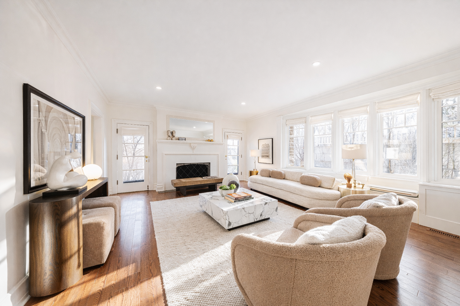 Bright living room with cream-colored sofas, a marble coffee table, large windows, hardwood floors, and a white fireplace.