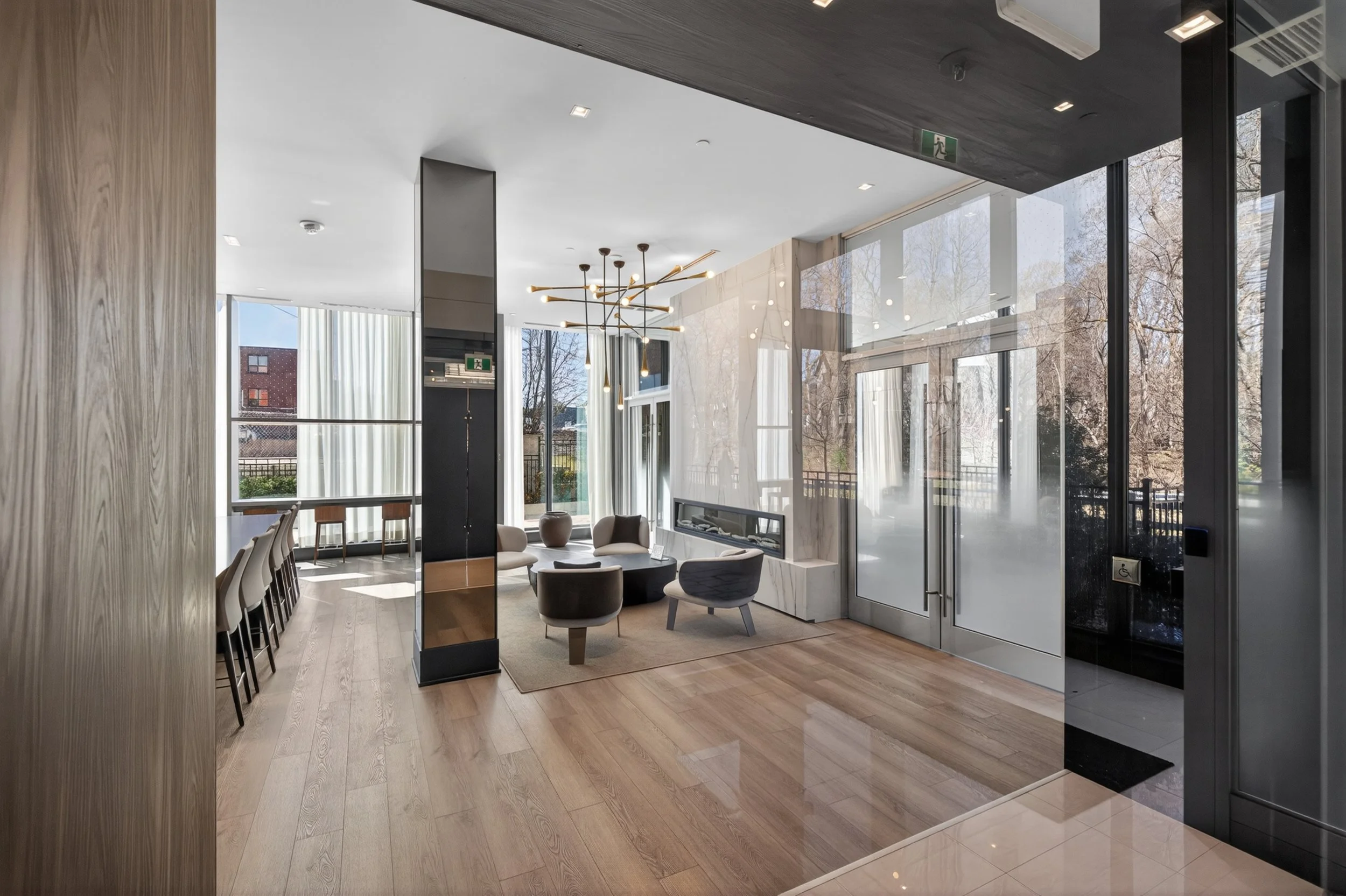 Modern hotel lobby with wood flooring, a seating area with black and white chairs, a marble fireplace, large windows letting in natural light, and a contemporary chandelier.