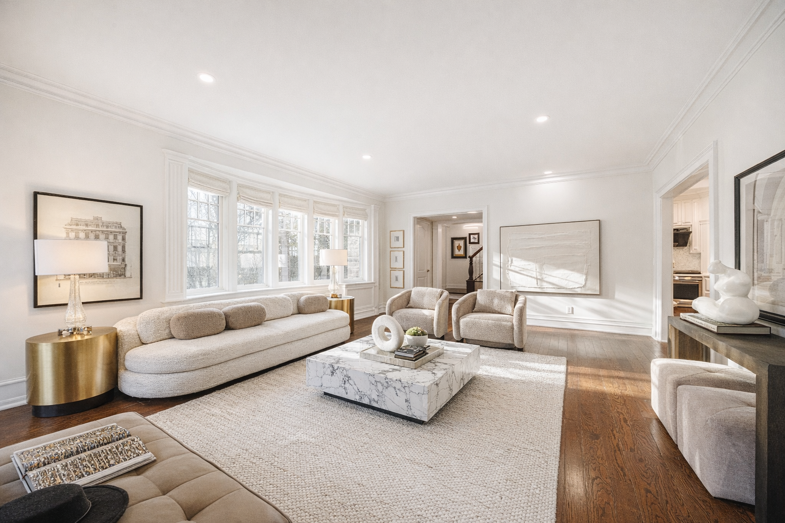 Bright living room with white walls, large window with multiple panes, beige sofa with rounded cushions, two beige armchairs, marble coffee table, wooden side tables with lamps, artwork on walls, and hardwood flooring.