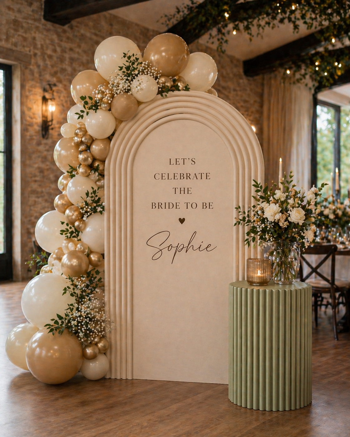 Decorative wedding or bridal shower display with a large beige arch with balloons and greenery on the left, and a green ribbed cylindrical table with a floral arrangement and candle on the right. The arch has the text "Let's celebrate the bride to be Sophie." The venue has exposed brick walls, large windows, and string lights.