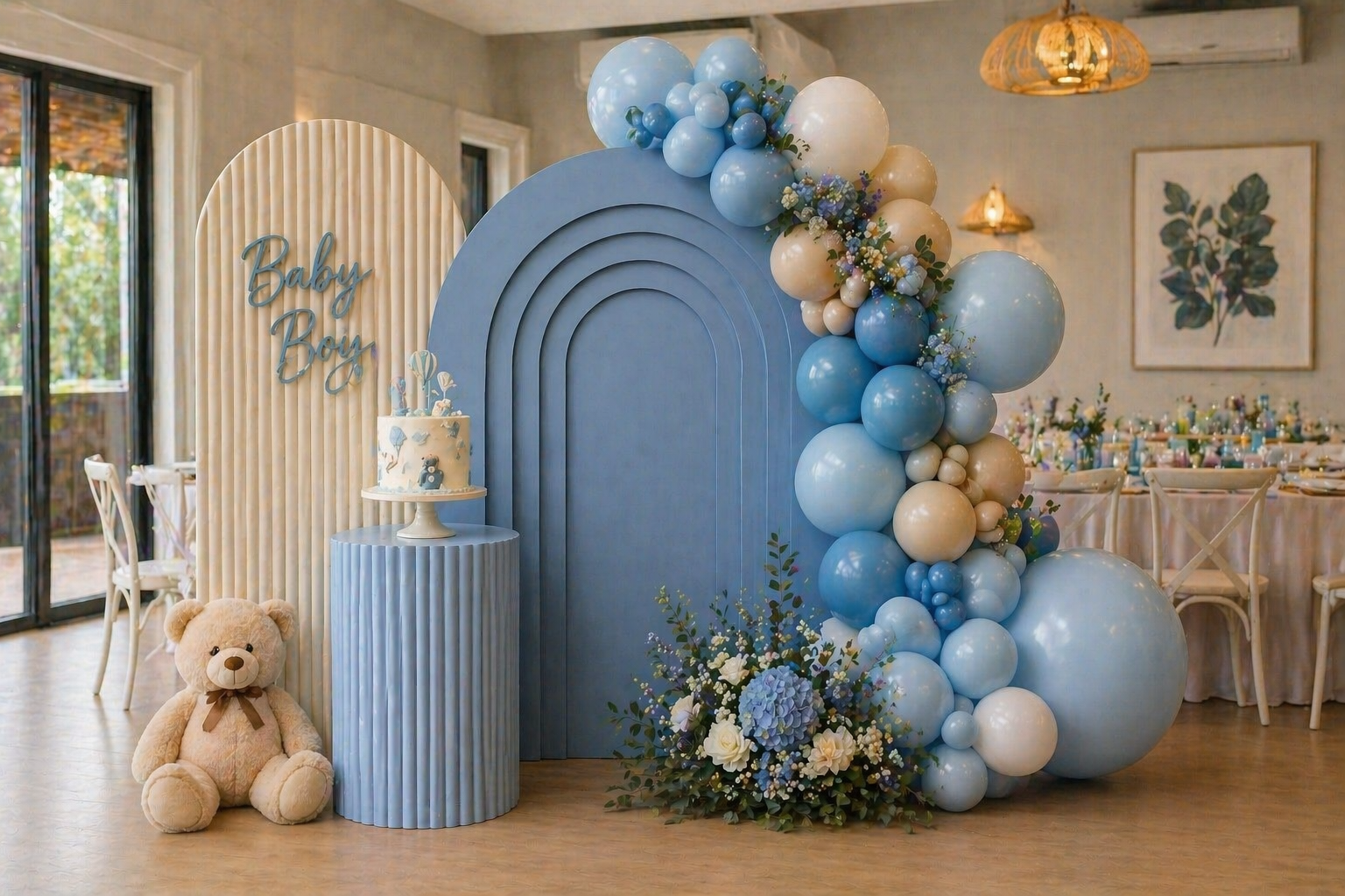 Decorative setup for a baby boy celebration featuring pastel blue and cream balloons, a teddy bear, a cake with stuffed teddy bear motif, and a sign that reads "Baby Boy" in a room with large windows and floral art on the walls.
