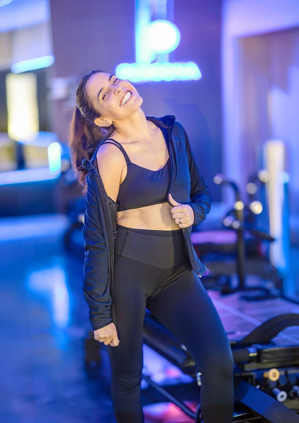 Smiling woman in a black jacket stands beside a Maven reformer under a glowing Persona Pilates logo