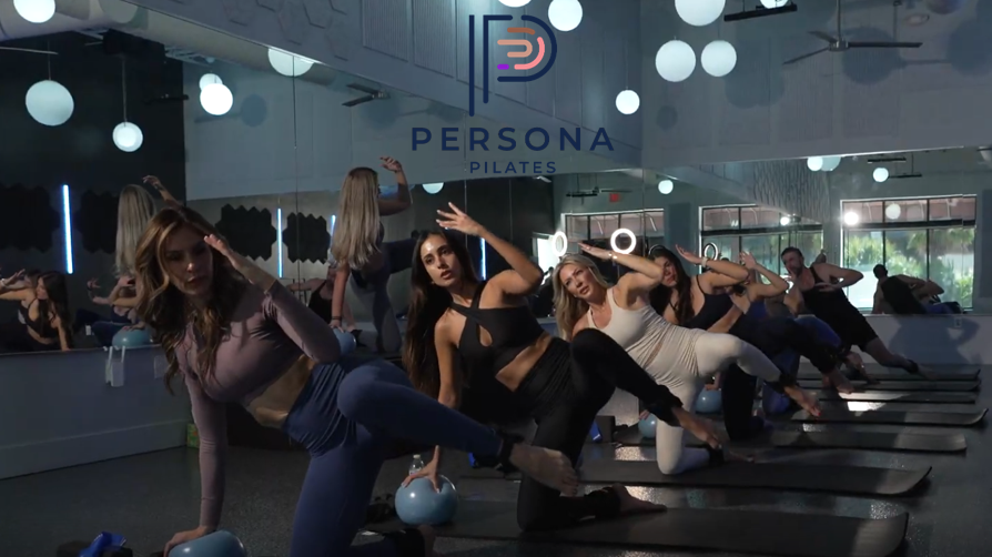 Women participating in a Pilates class at Perona Pilates studio, performing side-lying leg lifts on mats with small exercise balls nearby, mirrored wall with pendant lights overhead.