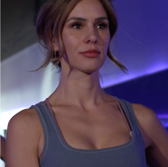 A woman with reddish-brown hair pulled back, wearing a sleeveless gray top, standing in a dimly lit environment with purple and white lighting.