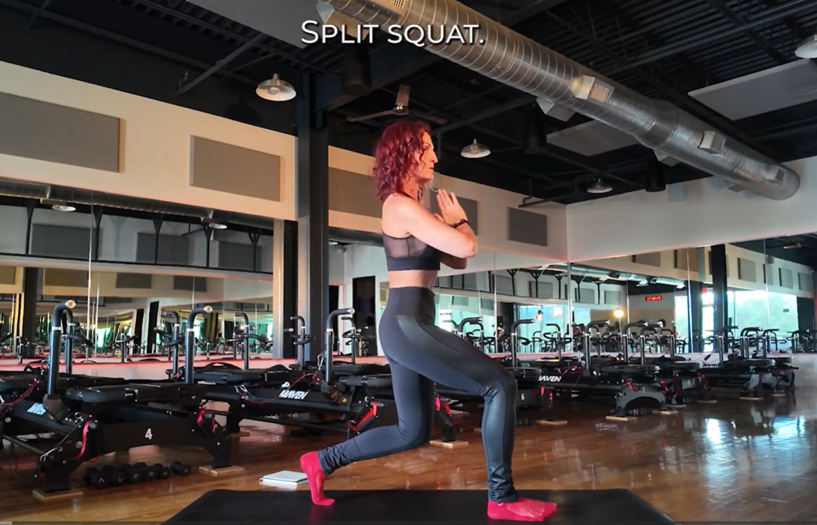 A woman in workout clothes performing a split squat exercise in a gym, with Mirrored walls and exercise equipment in the background.