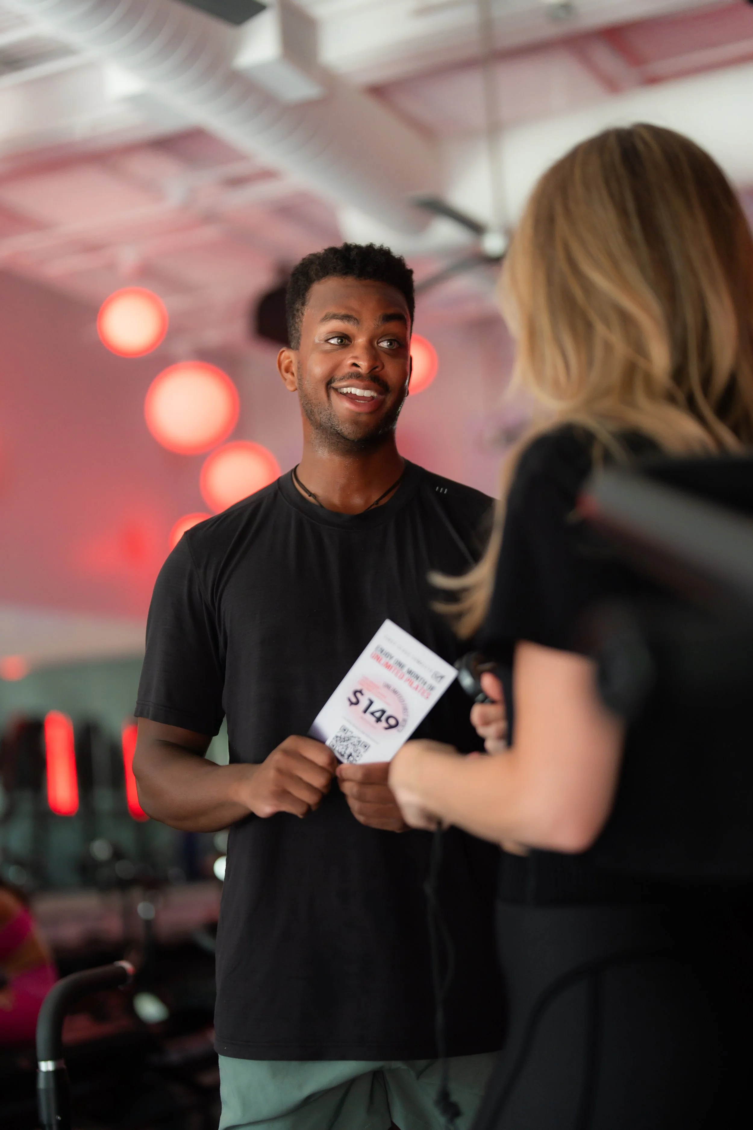 A young man smiling and talking to a woman, holding a ticket with $149 printed on it, in a room with pink and red lighting and hanging round lights.