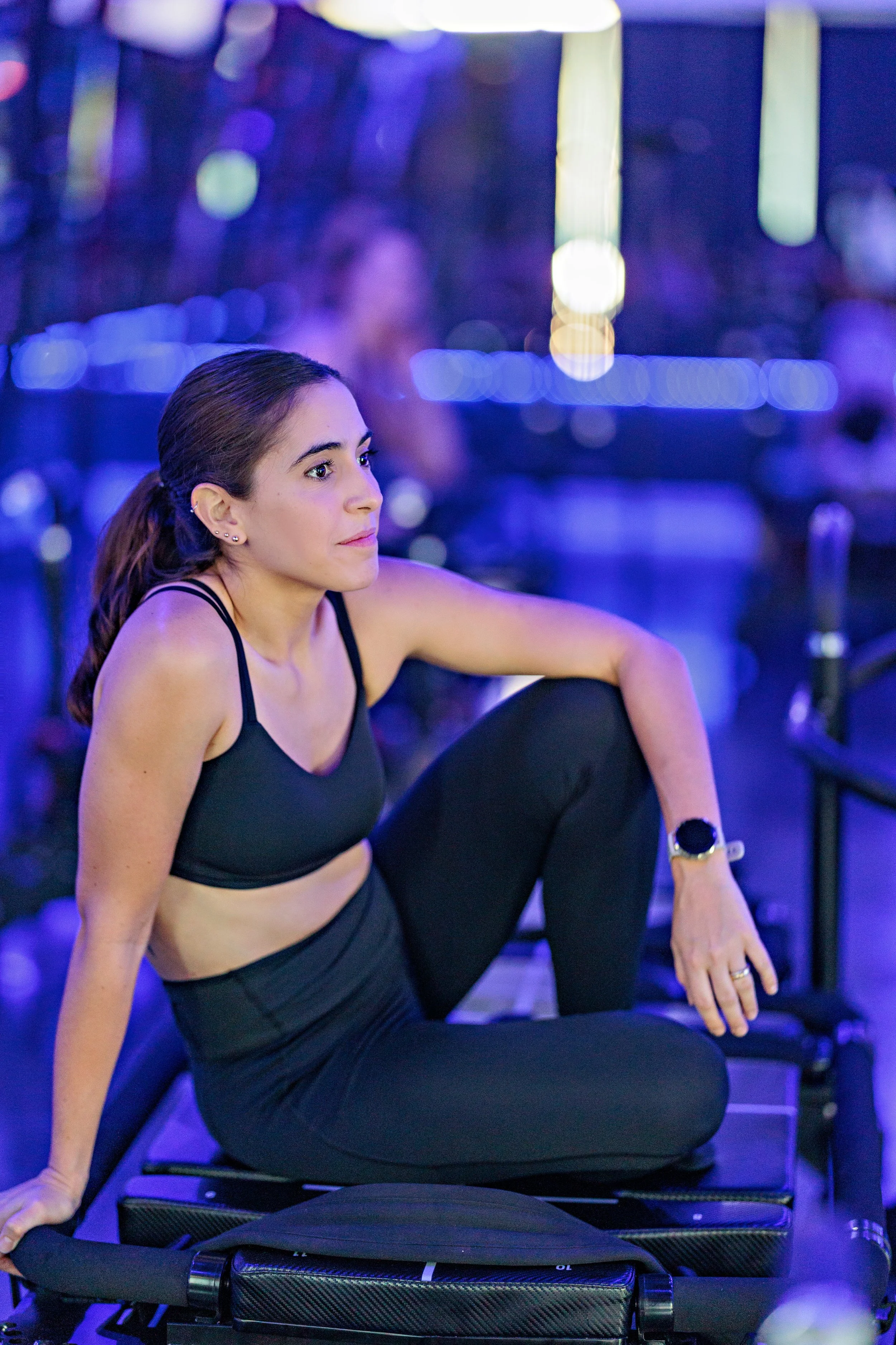 Young woman in black athletic wear, sitting on a bench in a gym, stretching her leg and looking to the side with a focused expression, with blue and purple gym lighting in the background.