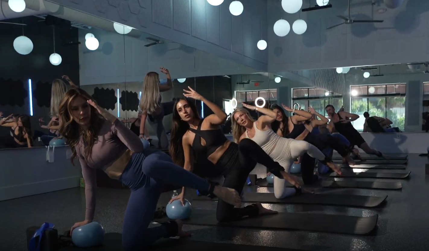Women participating in a fitness or yoga class in a mirrored studio, balancing on one knee with their hands raised near their heads, dressed in activewear.