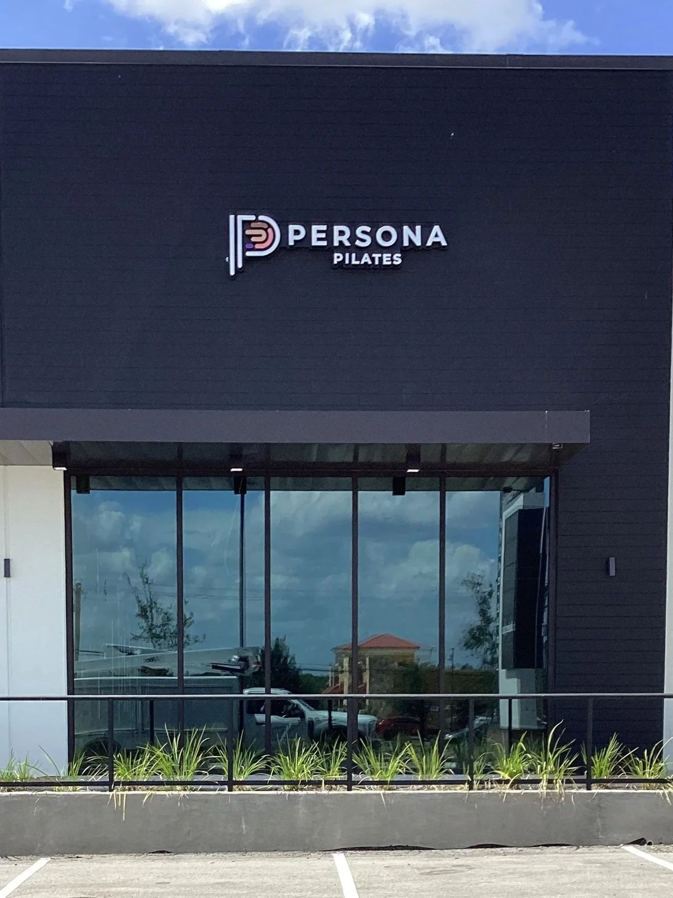 Exterior of a building with a sign that reads 'Persona Pilates,' featuring a black wall, large glass windows, and a parking lot in the foreground.