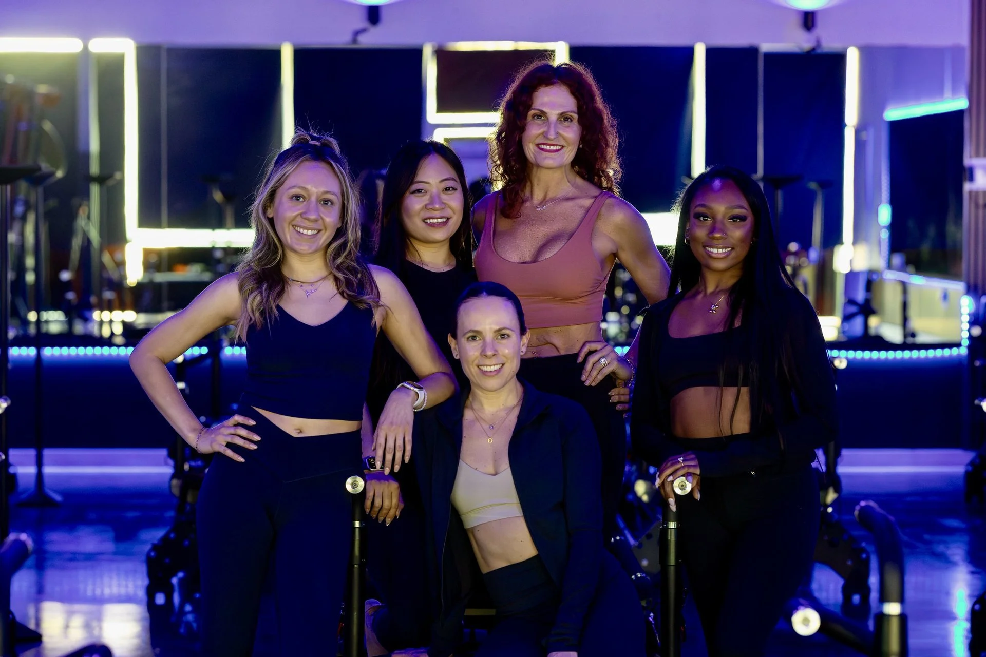 Five Persona Pilates instructors pose together among Maven reformers in a blue-lit studio