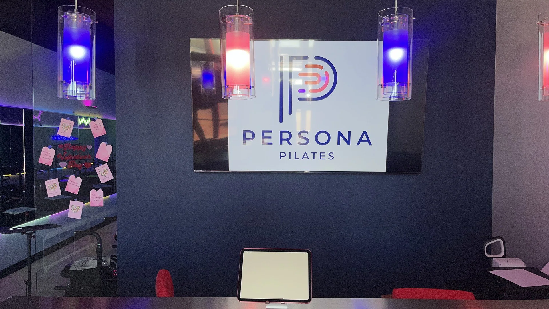 Persona Pilates check-in desk with glowing logo sign, tablet, and neon pendant lights in an empty lobby
